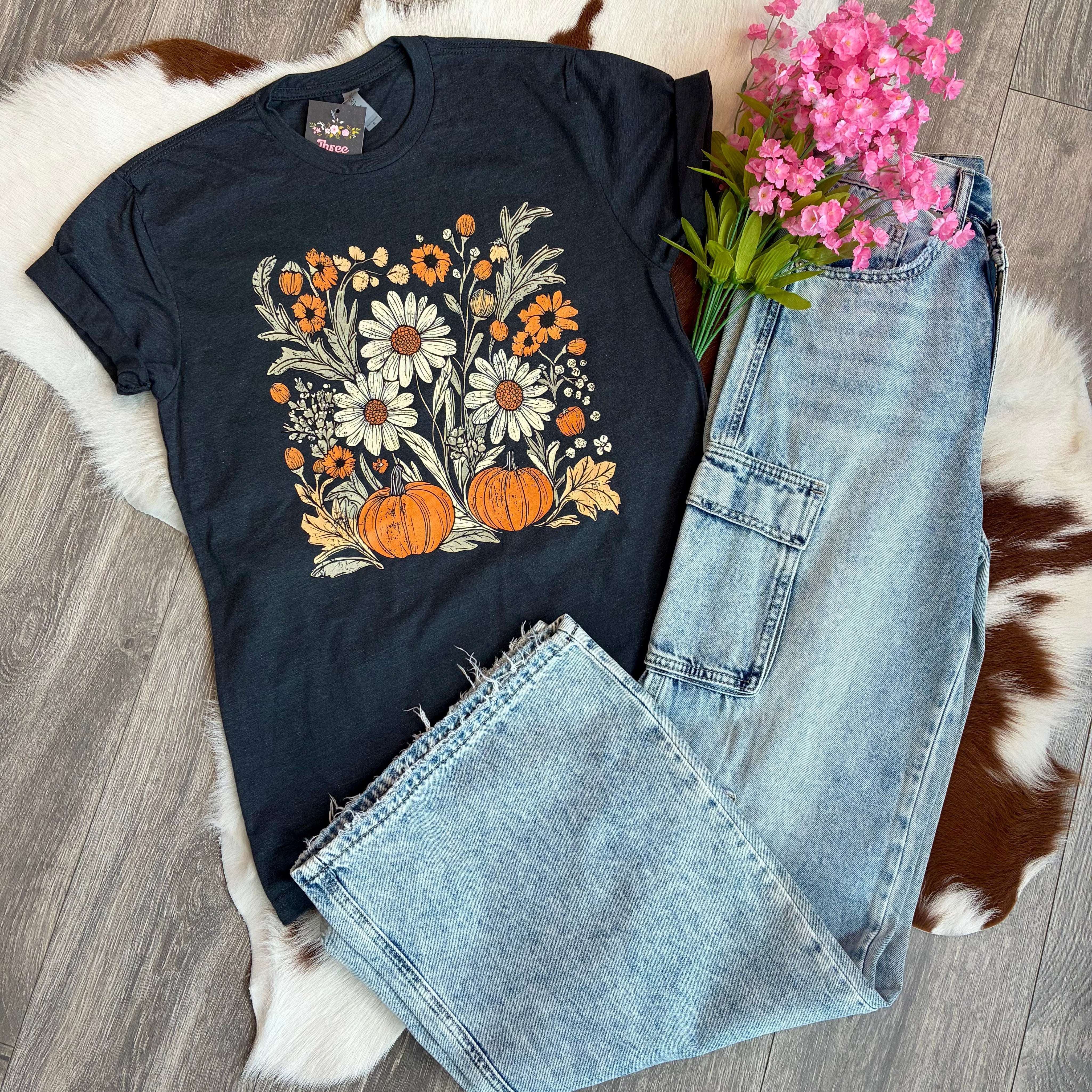 Black t-shirt with floral and pumpkin design, paired with jeans and flowers on a wooden floor.