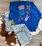Blue sweatshirt with colorful designs on a wooden floor with denim shorts and a plant.