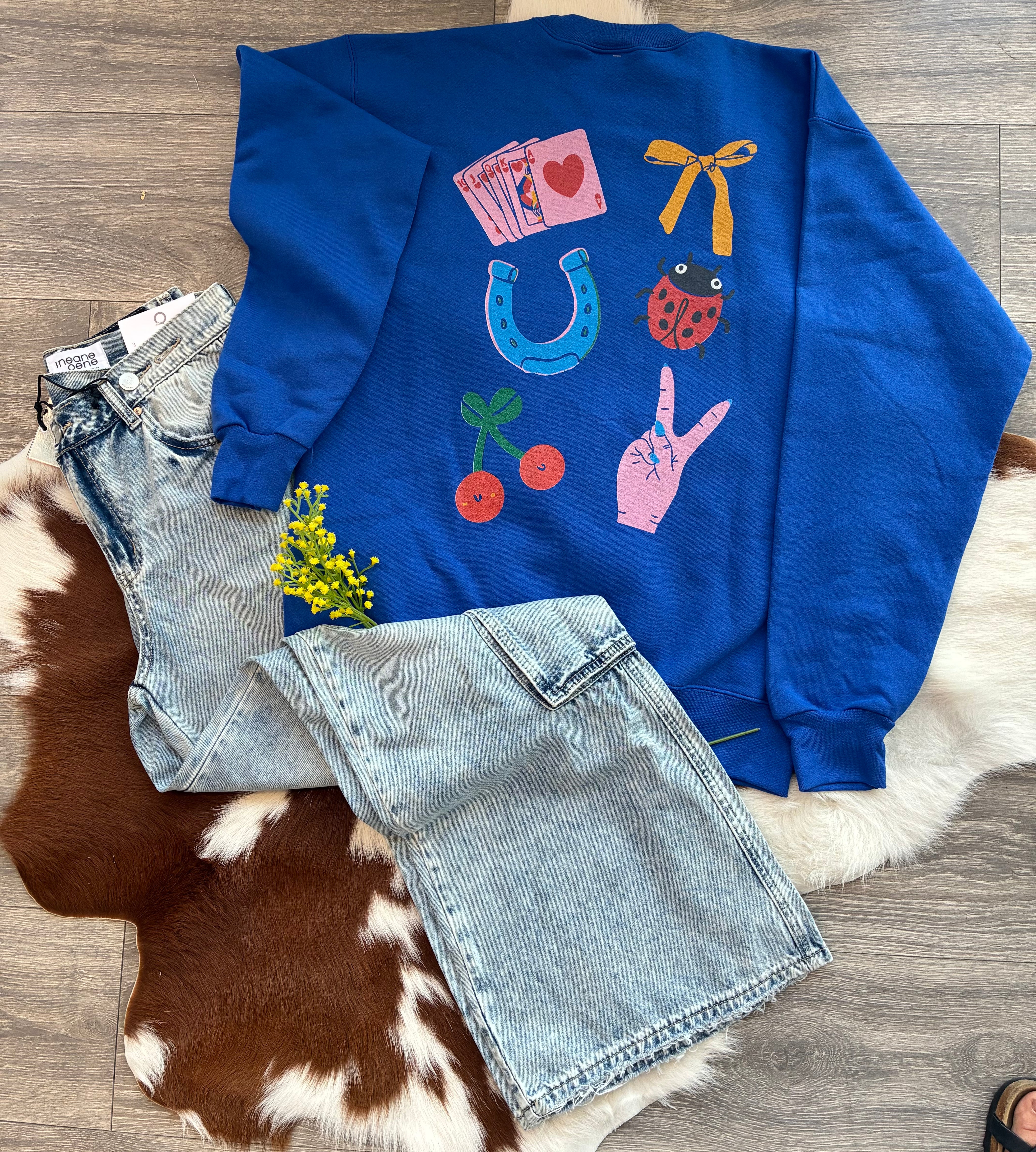 Blue sweatshirt with colorful designs on a wooden floor with denim shorts and a plant.