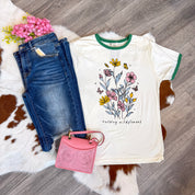 Flat lay of a casual boutique outfit featuring a cream graphic tee with green trim and a floral design that reads “raising wildflowers,” paired with distressed dark wash denim jeans, a tooled pink mini handbag, and a bunch of pink faux flowers, all arranged on a cowhide rug over wood flooring.