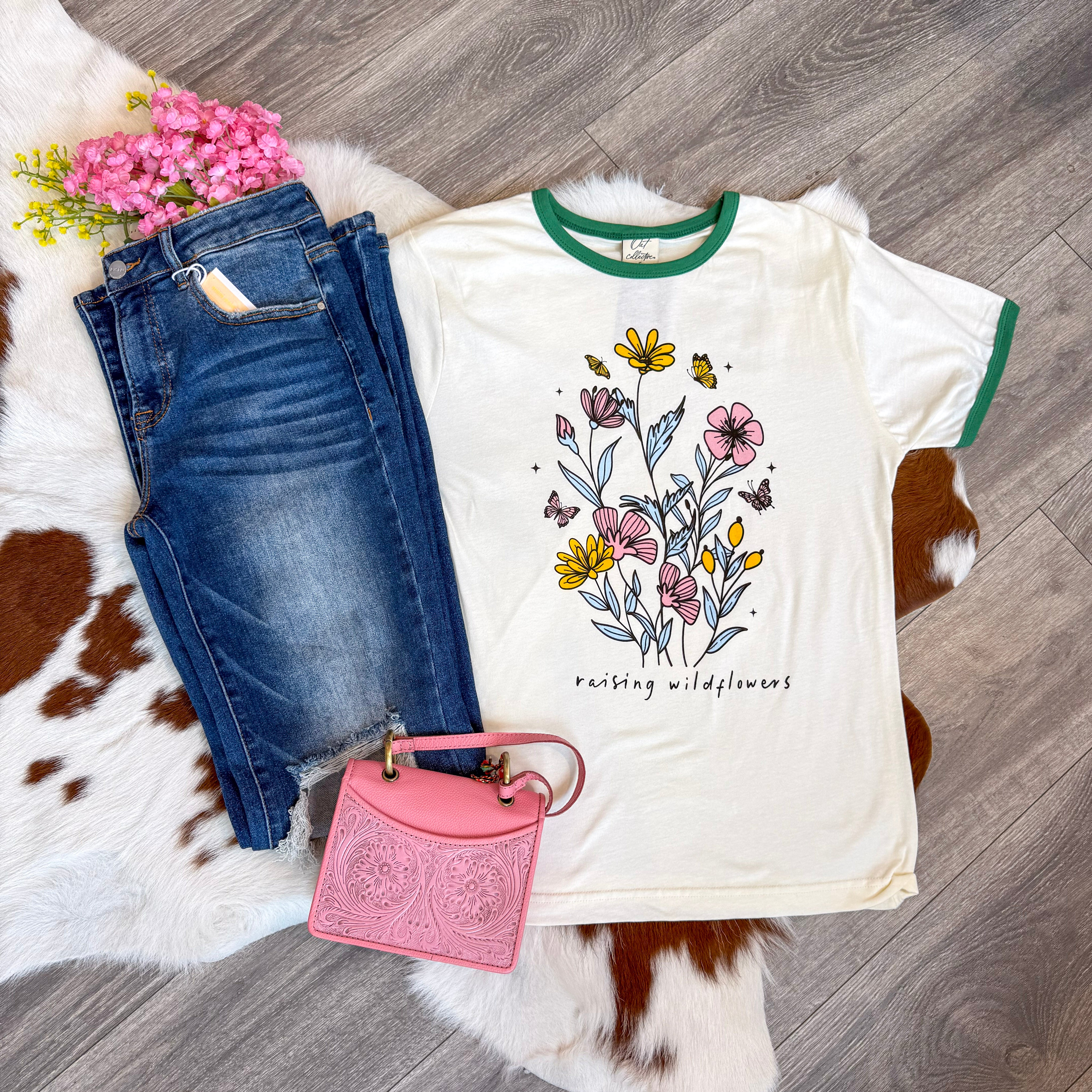 Flat lay of a casual boutique outfit featuring a cream graphic tee with green trim and a floral design that reads “raising wildflowers,” paired with distressed dark wash denim jeans, a tooled pink mini handbag, and a bunch of pink faux flowers, all arranged on a cowhide rug over wood flooring.