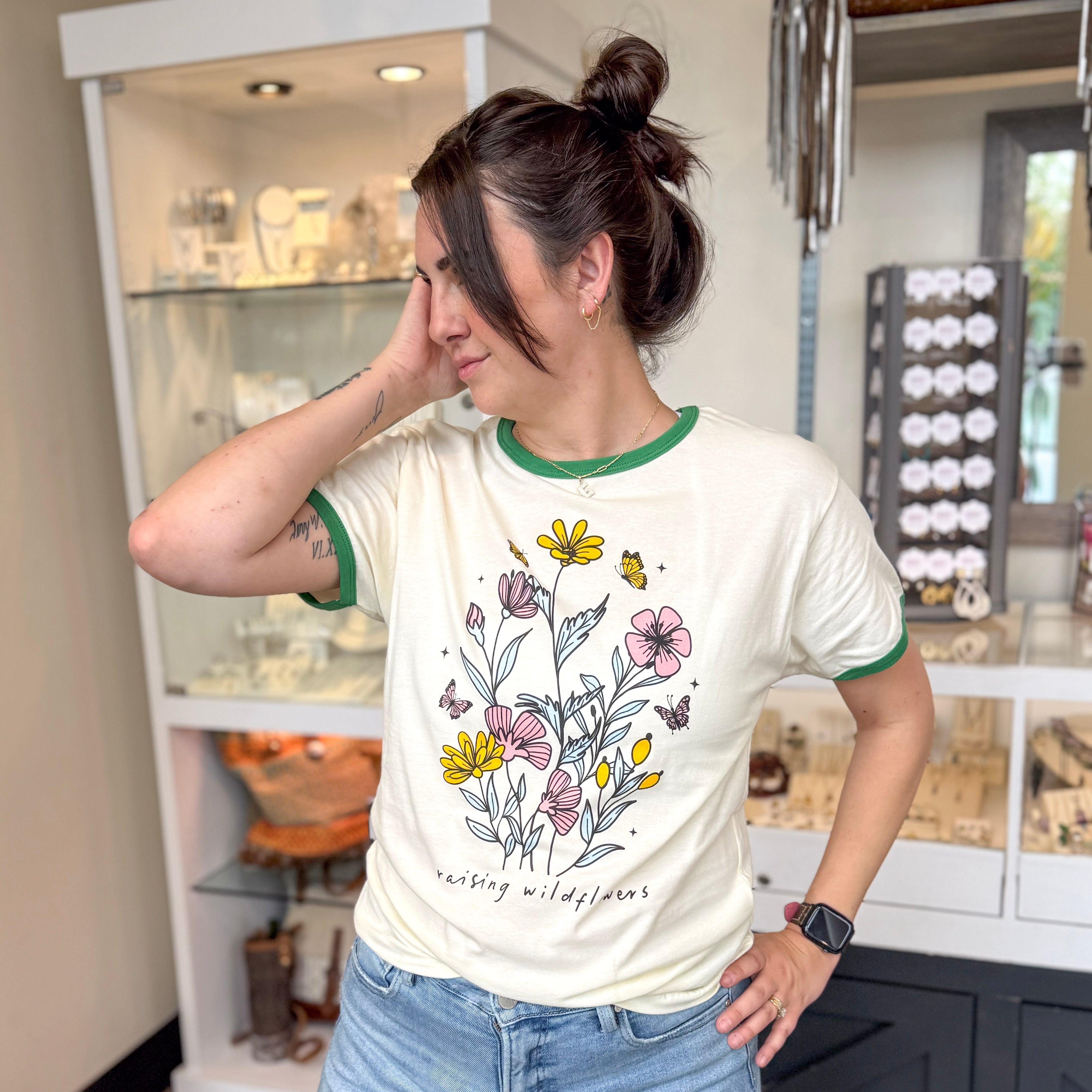 Woman posing inside a boutique wearing a cream graphic tee with green trim that features a colorful wildflower bouquet and the phrase “raising wildflowers,” styled with light wash jeans, gold jewelry, and an Apple Watch, with a jewelry display case in the background.
