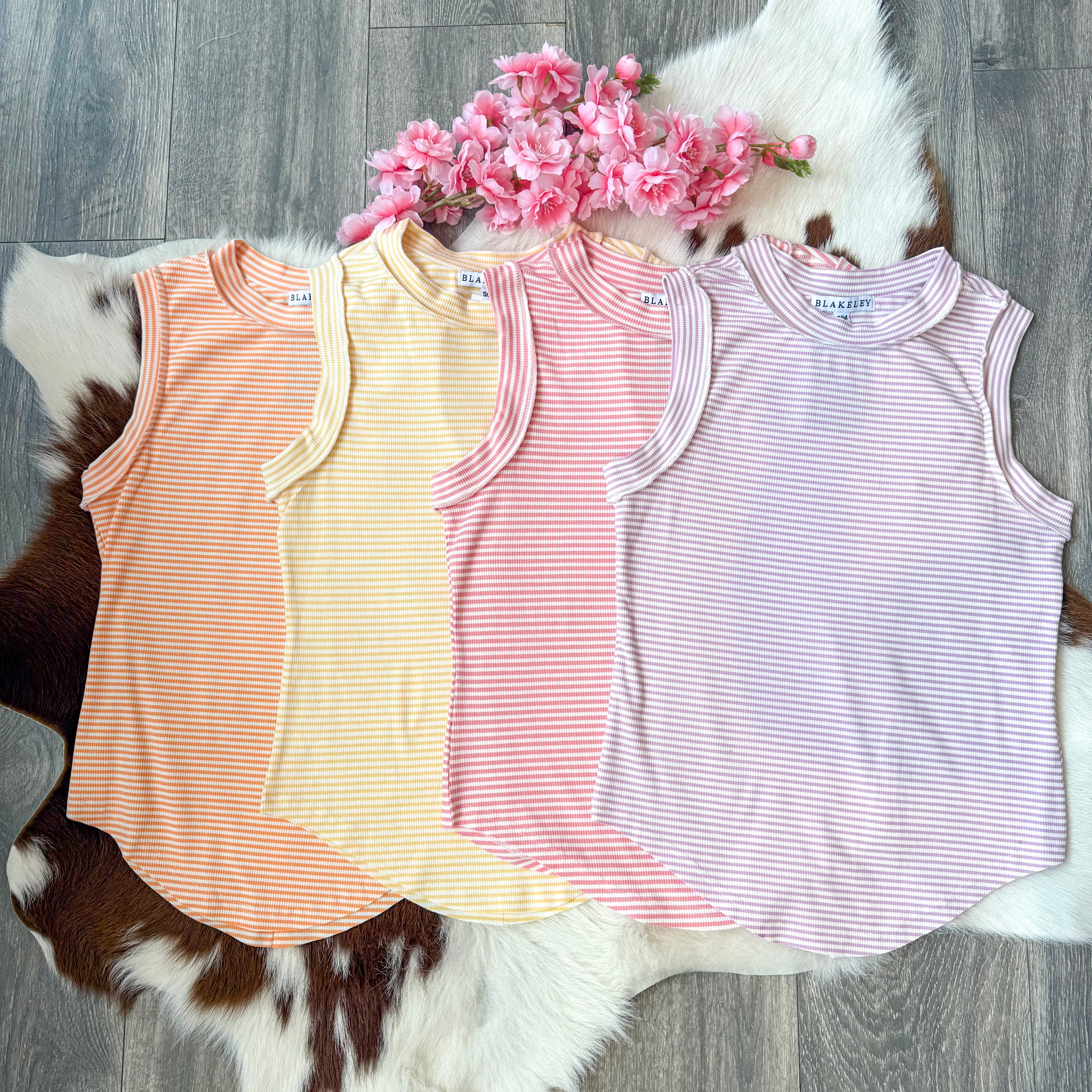 Flat lay of four adult-sized ribbed tank tops in pastel striped colors—orange, yellow, pink, and lavender—arranged side by side on a cowhide rug with pink faux flowers placed above, displayed on a wood floor background.
