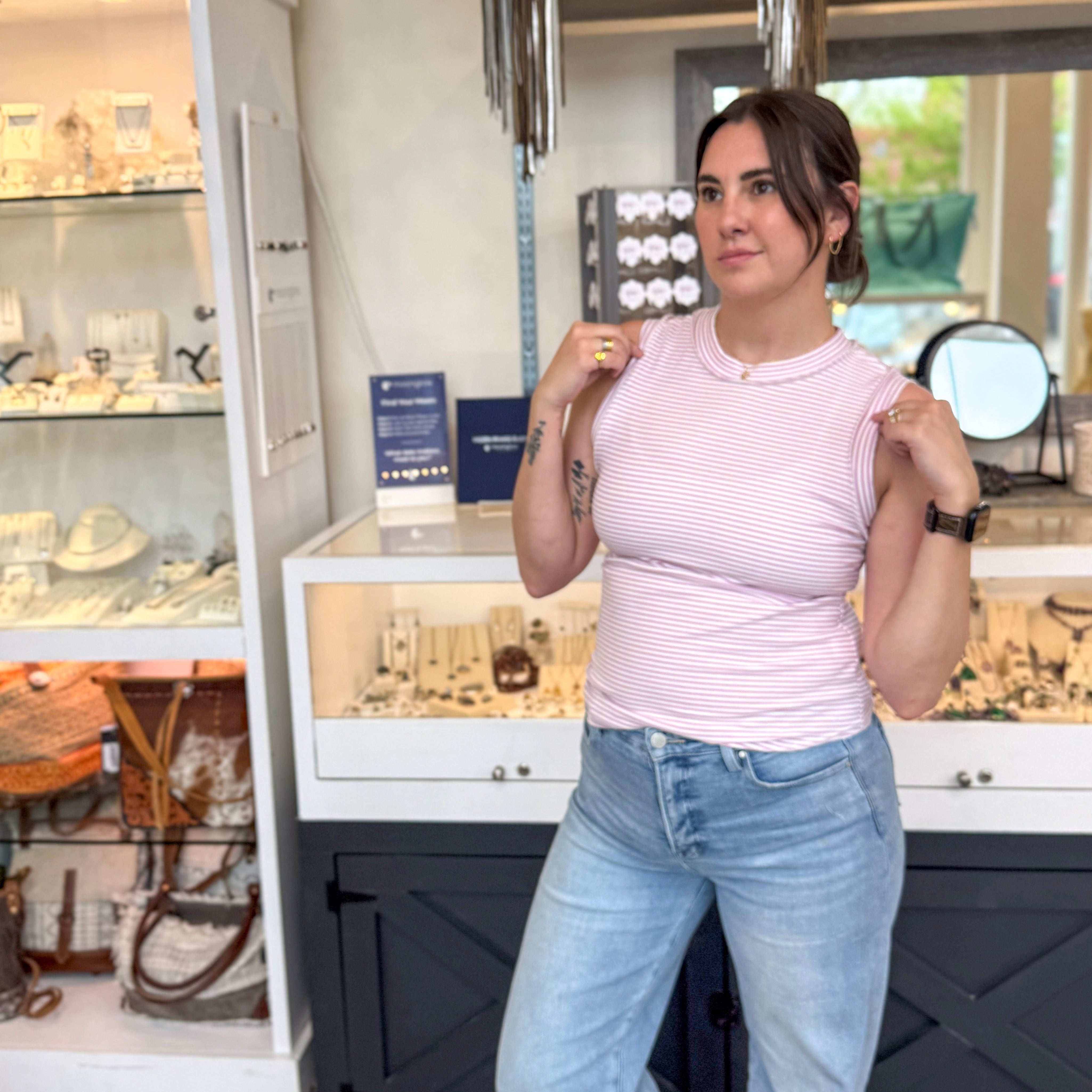 Woman posing inside a boutique wearing a fitted lavender and white striped ribbed tank top, styled with light wash high-waisted jeans, gold jewelry, and an Apple Watch, standing in front of a glass jewelry display case.