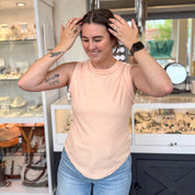 Woman smiling and adjusting her hair while wearing a peach and white striped ribbed tank top, styled with light wash jeans, gold rings, and an Apple Watch, standing inside a boutique in front of a jewelry and accessories display.