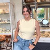 Woman smiling and posing inside a boutique while wearing a yellow and white striped ribbed tank top, tucked into high-waisted light wash jeans, accessorized with an Apple Watch, gold rings, and hoop earrings, with jewelry displays visible in the background.