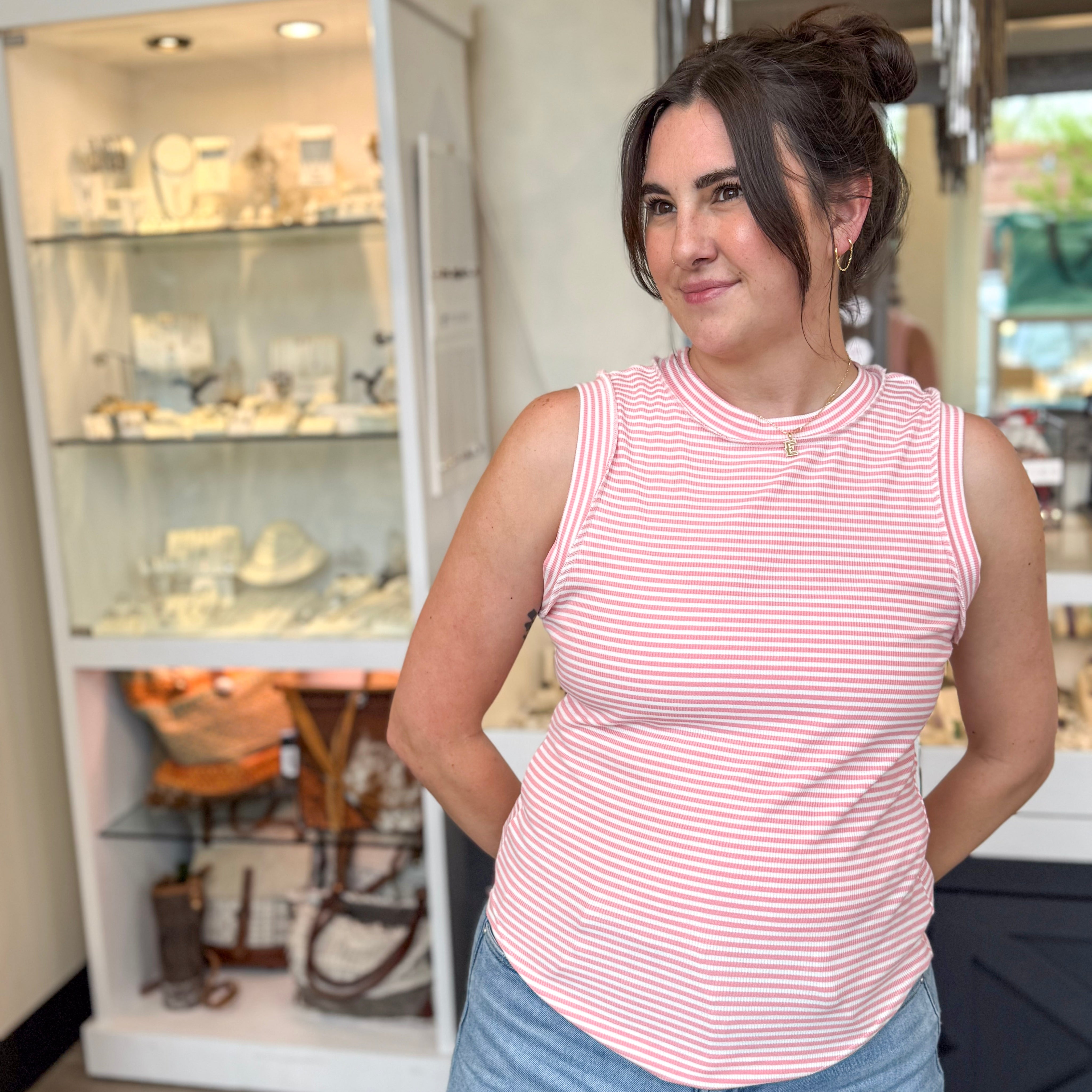 Woman posing inside a boutique wearing a pink and white striped ribbed tank top, styled with light wash jeans, gold hoop earrings, and a delicate initial necklace, standing in front of a jewelry display case.