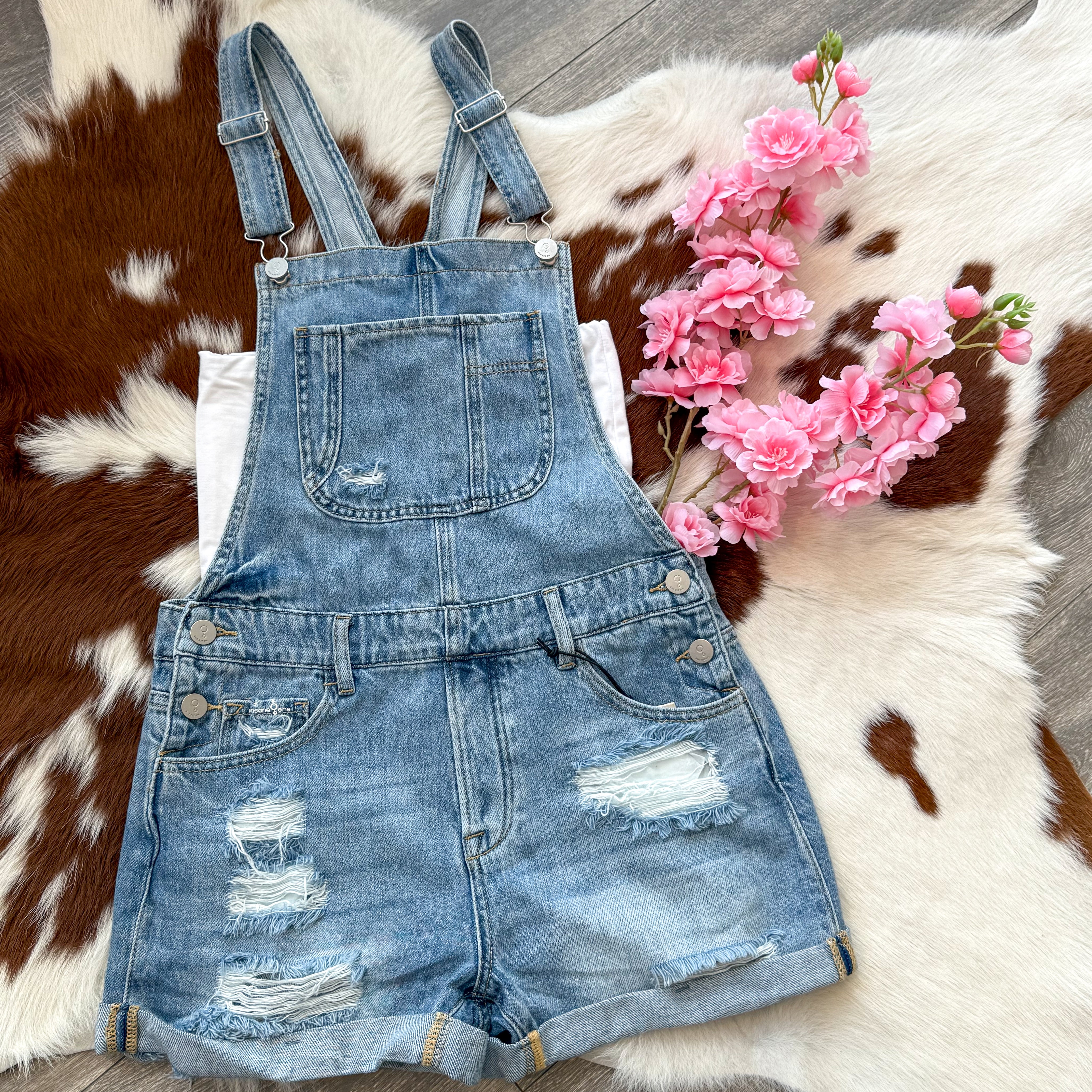 Flat lay of distressed denim short overalls with rolled cuffs, styled over a white shirt and placed on a cowhide rug with pink flowers for a rustic, summery vibe.
