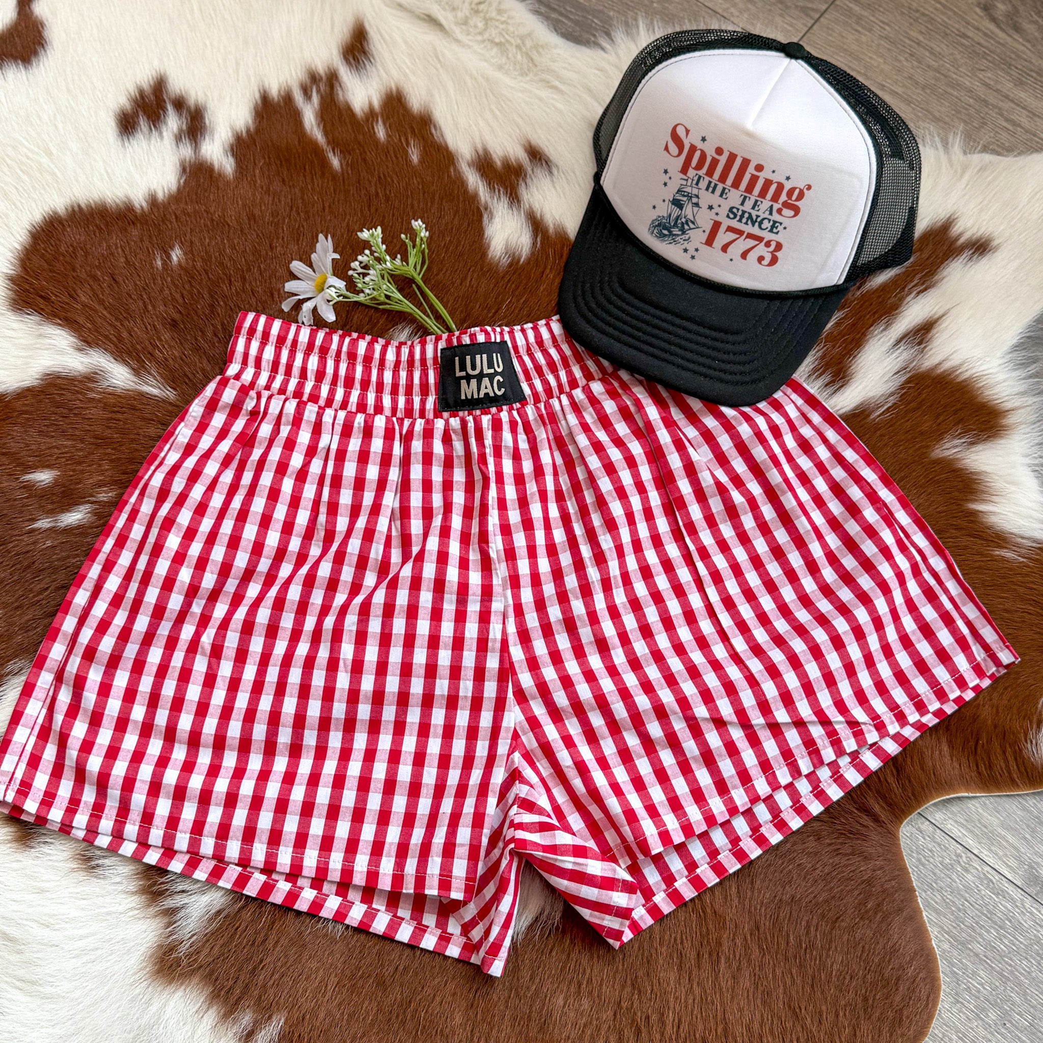 Flat lay of red and white gingham shorts with an elastic waistband and "Lulu Mac" label, styled on a cowhide rug next to a black-and-white trucker hat that reads "Spilling the Tea Since 1773," with white daisies placed beside the shorts – a festive and playful Fourth of July look.