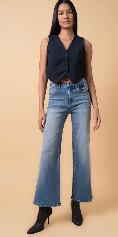 Woman wearing high-rise flare leg jeans in a medium blue wash, styled with a navy vest and black heeled boots against a tan background.
