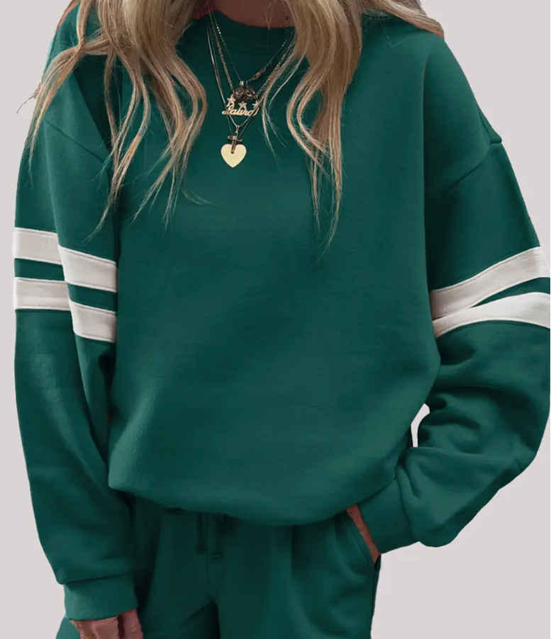 Woman wearing a dark green oversized pullover sweatshirt with white varsity-style sleeve stripes, paired with matching shorts and layered gold necklaces.