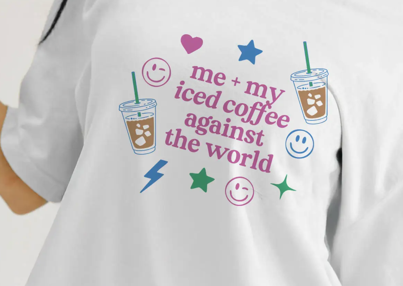 Close-up of a white graphic t-shirt featuring the phrase 'me + my iced coffee against the world' along with colorful illustrations of iced coffee cups, smiley faces, and stars.