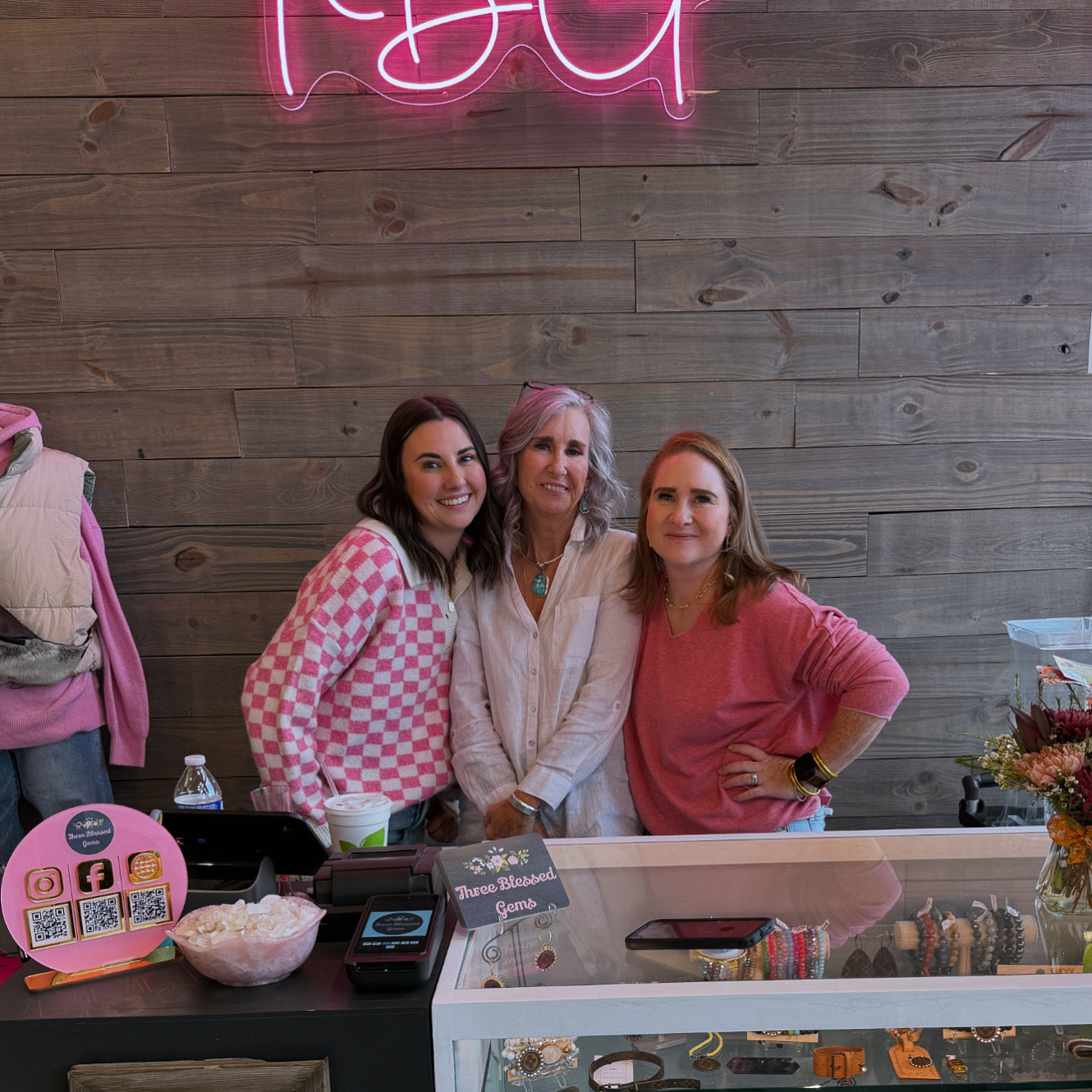 Three women smiling behind the checkout counter at Three Blessed Gems boutique. They are dressed in stylish pink and neutral outfits, standing in front of a rustic wooden wall with a glowing pink neon sign. The boutique’s counter displays jewelry, a sign with the store’s name, a QR code stand for social media, and a bowl of decorative stones. The cozy and welcoming atmosphere reflects the boutique’s fun and friendly vibe.