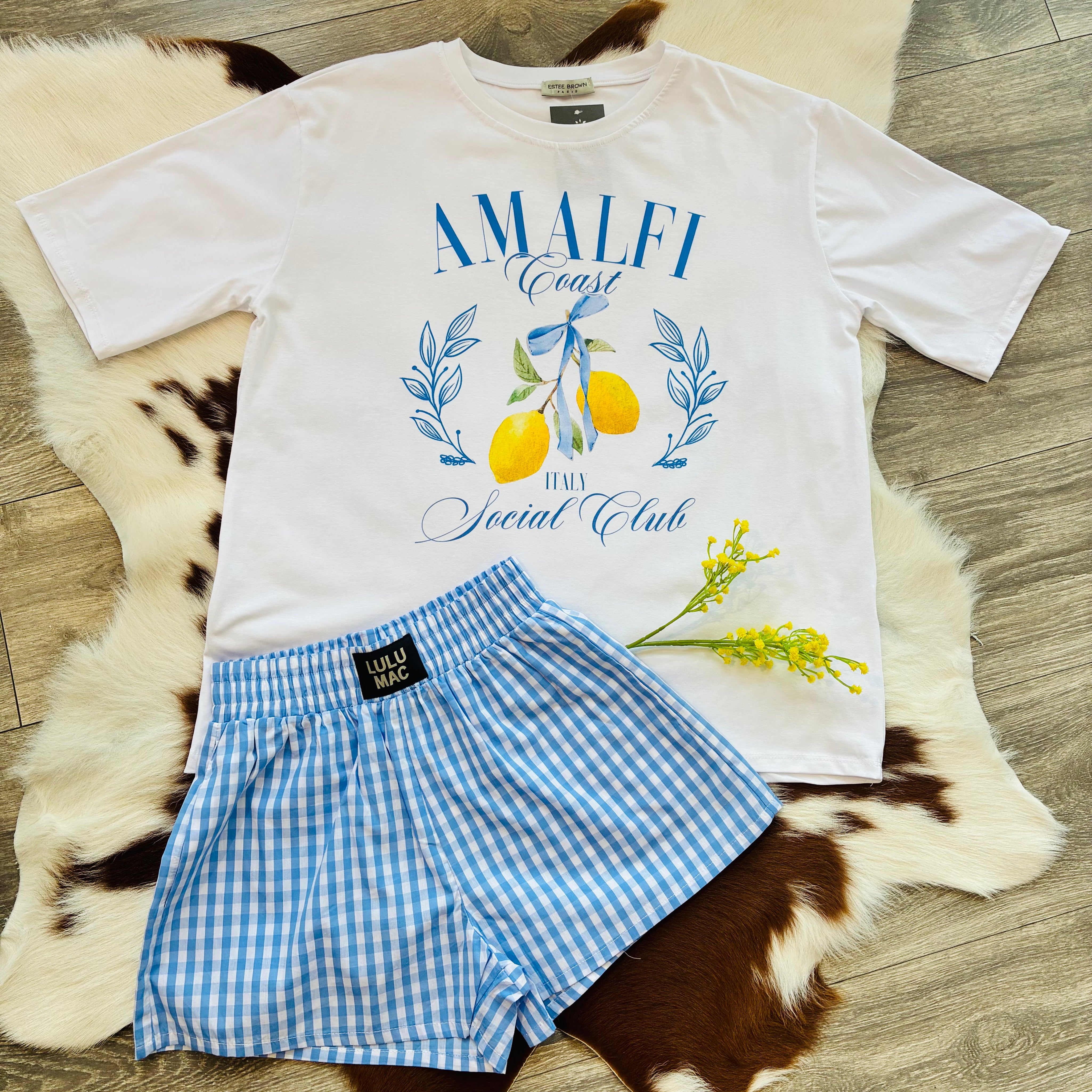 Flat lay of a light beige sweatshirt with a “Lemon Market, Positano Italy” graphic featuring illustrated lemons, leaves, and text, styled next to a pair of dark-wash jeans and yellow flowers on a faux fur and cowhide rug over wooden flooring.