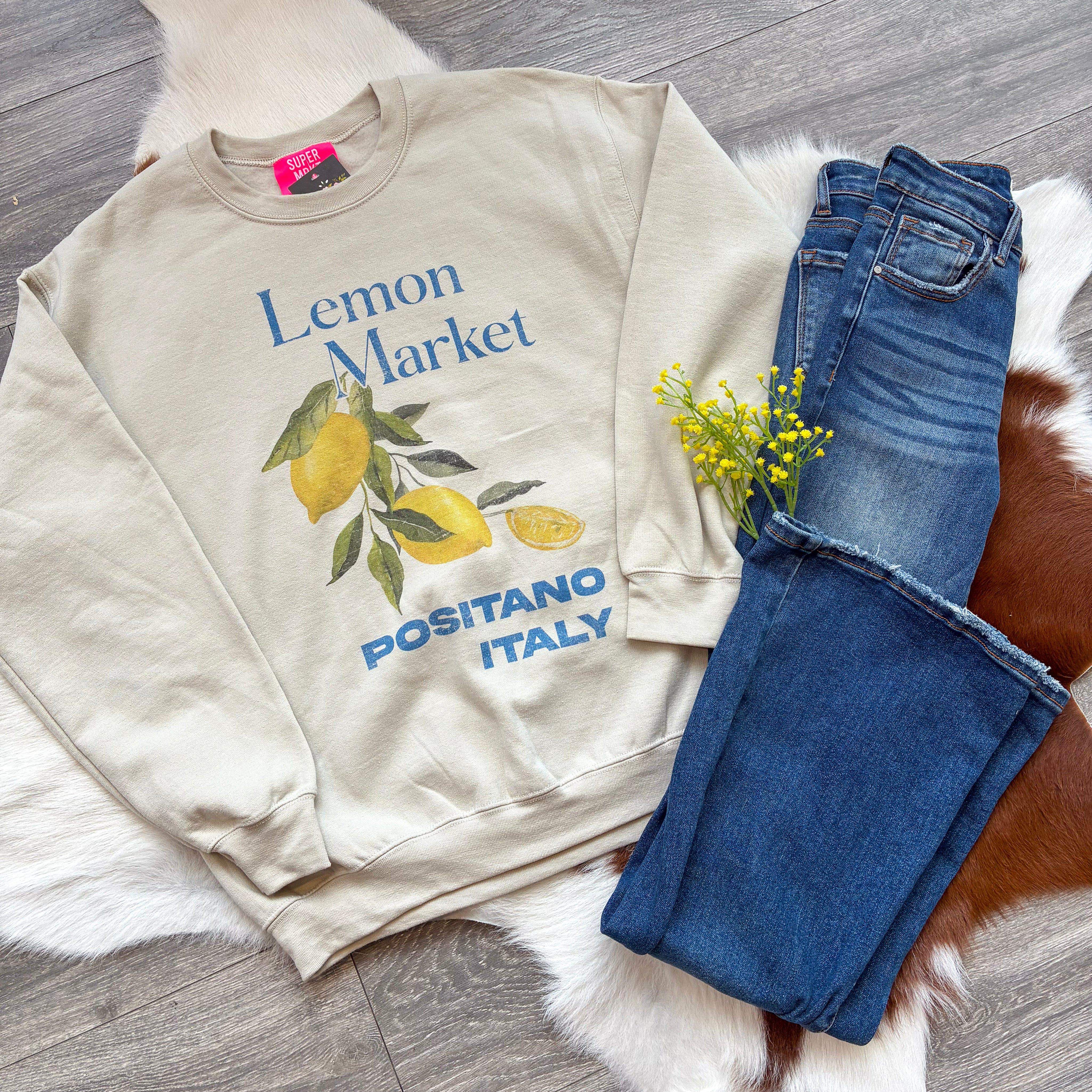  Flat-lay of a beige sweatshirt with a “Lemon Market, Positano Italy” graphic featuring watercolor-style lemons and greenery, styled next to dark-wash jeans and a sprig of yellow flowers on a cowhide and faux fur rug over wood flooring.