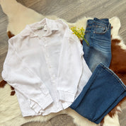 Flat lay of a white textured gauze button-up shirt paired with dark-wash jeans and a small yellow floral sprig, displayed on a white and brown faux fur rug with a wooden floor background.