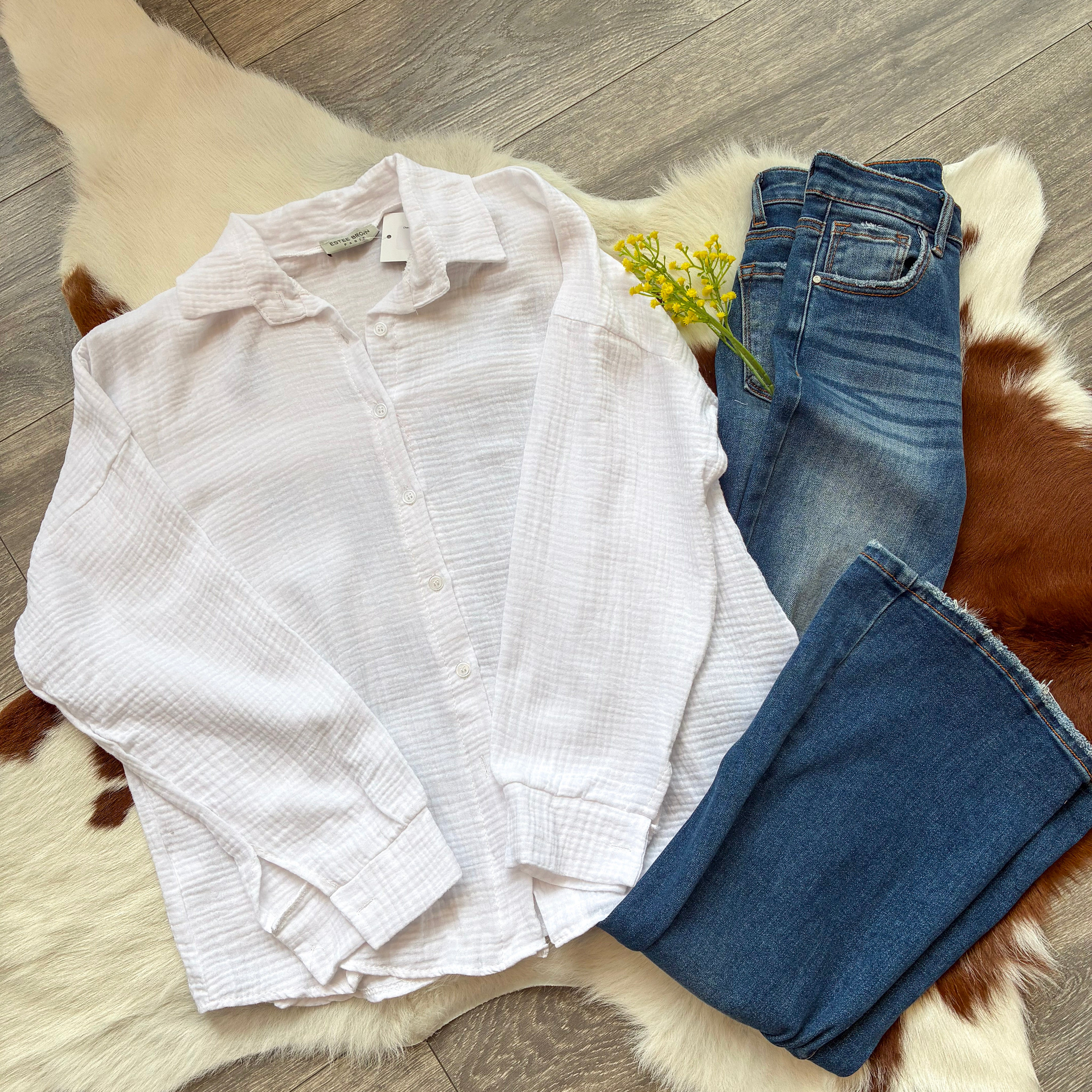 Flat lay of a white textured gauze button-up shirt paired with dark-wash jeans and a small yellow floral sprig, displayed on a white and brown faux fur rug with a wooden floor background.