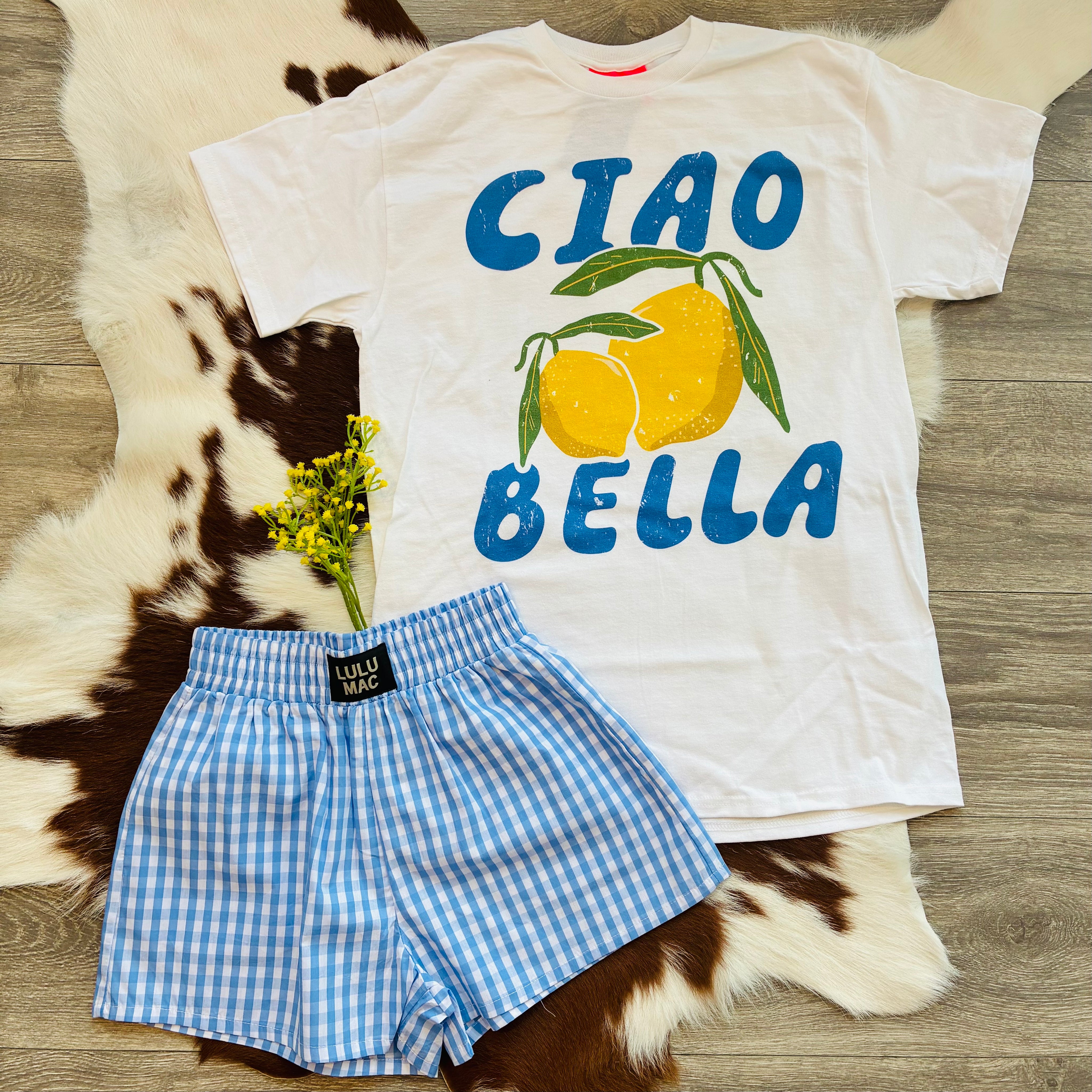 White t-shirt with 'CIAO BELLA' and lemon design, paired with blue checkered shorts on a wooden surface.