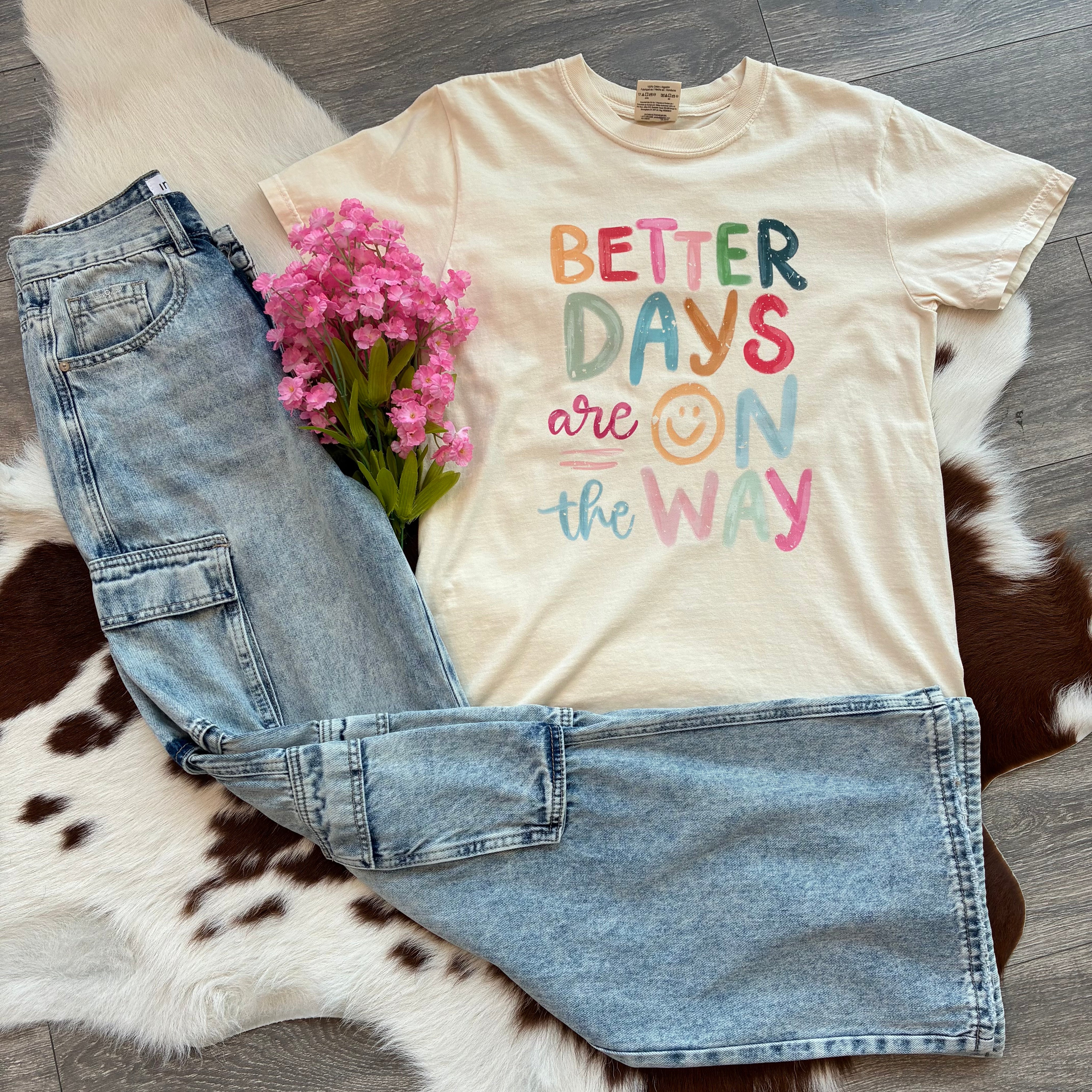 Cream graphic tee with the phrase "Better Days Are On The Way" in colorful letters, styled with light wash cargo jeans and pink flowers, laid flat on a cowhide rug.
