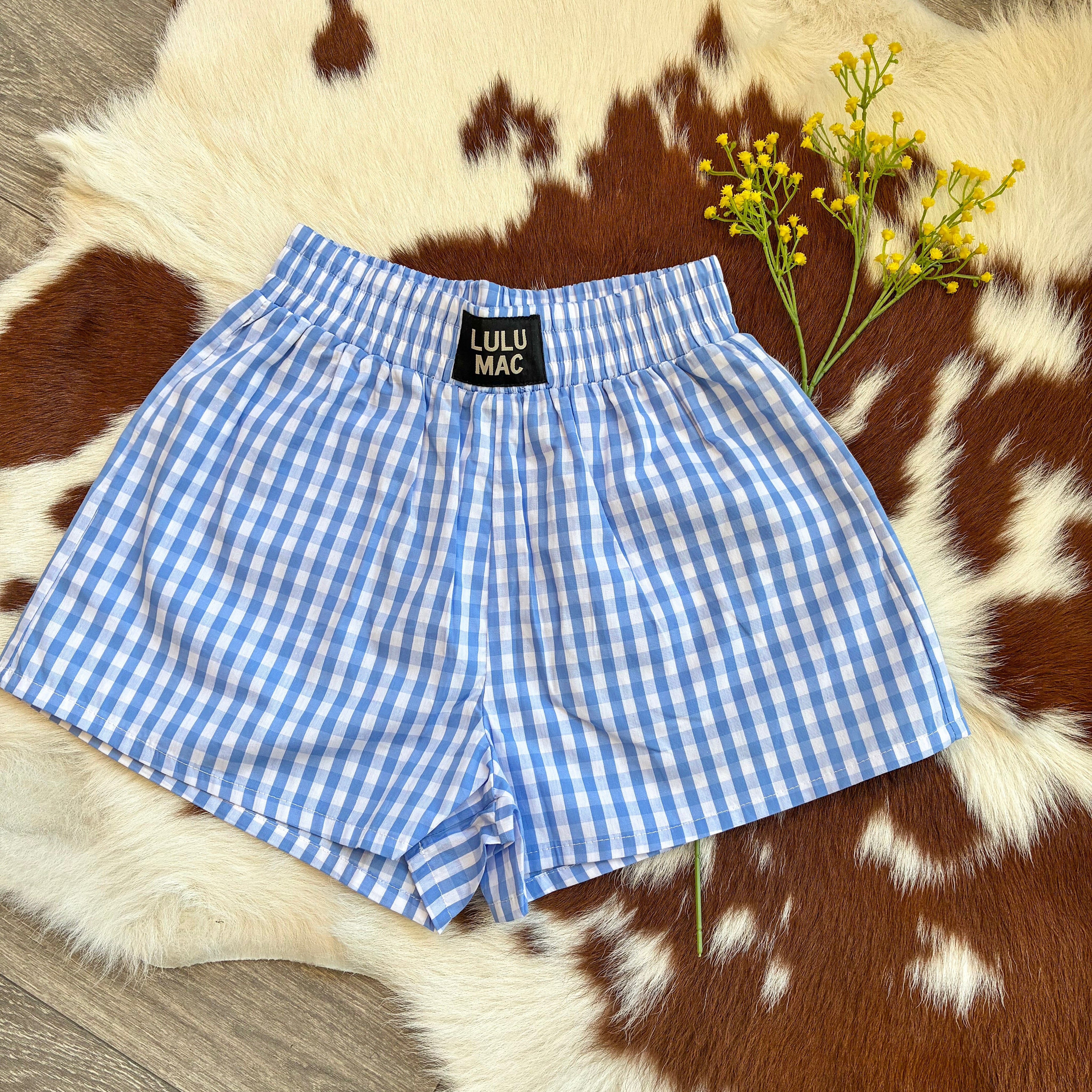 Blue gingham Lulu Mac lounge shorts displayed on a brown and white cowhide rug with yellow flowers, styled flat lay for boutique summer fashion.