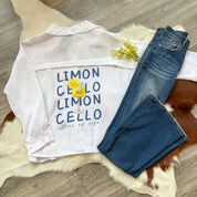 Flat lay of a white gauze button-up shirt with a playful “LIMONCELLO” graphic and lemon drink illustration, styled with dark-wash jeans and yellow flowers on a faux fur and cowhide rug.