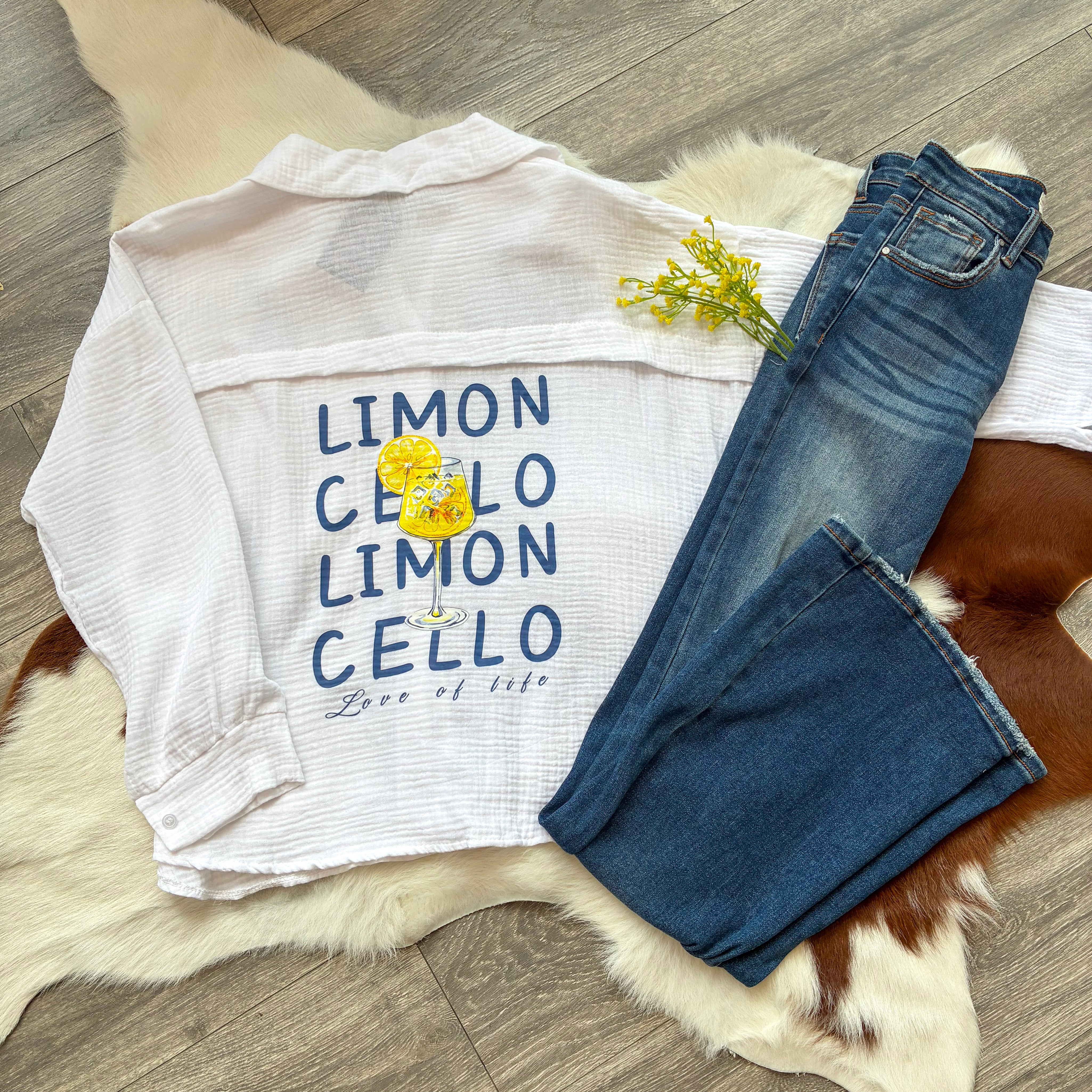 Flat lay of a white gauze button-up shirt with a playful “LIMONCELLO” graphic and lemon drink illustration, styled with dark-wash jeans and yellow flowers on a faux fur and cowhide rug.