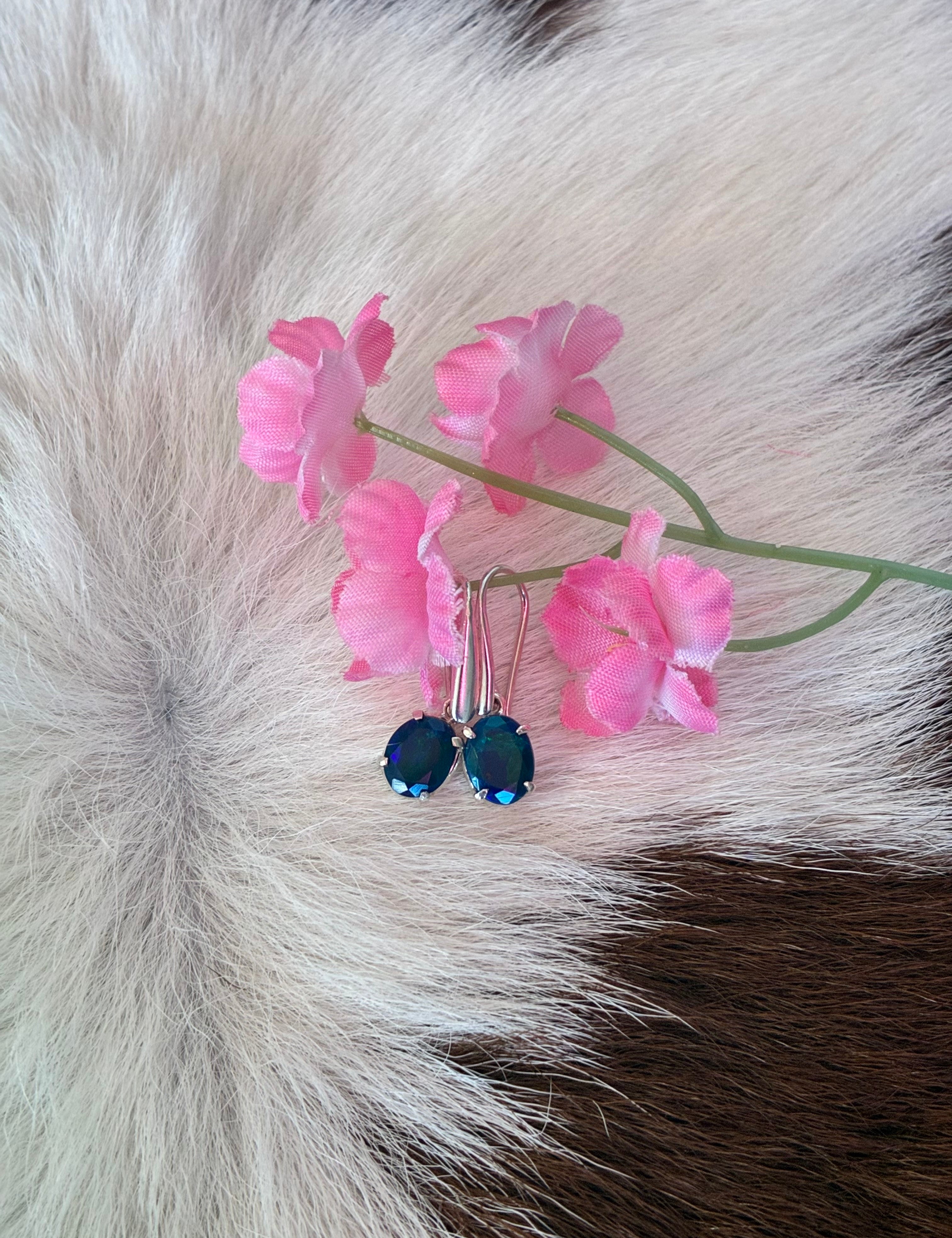 Black Ethiopian opal earrings with vibrant green and blue hues set in silver, placed on a textured fur background with pink floral accents.