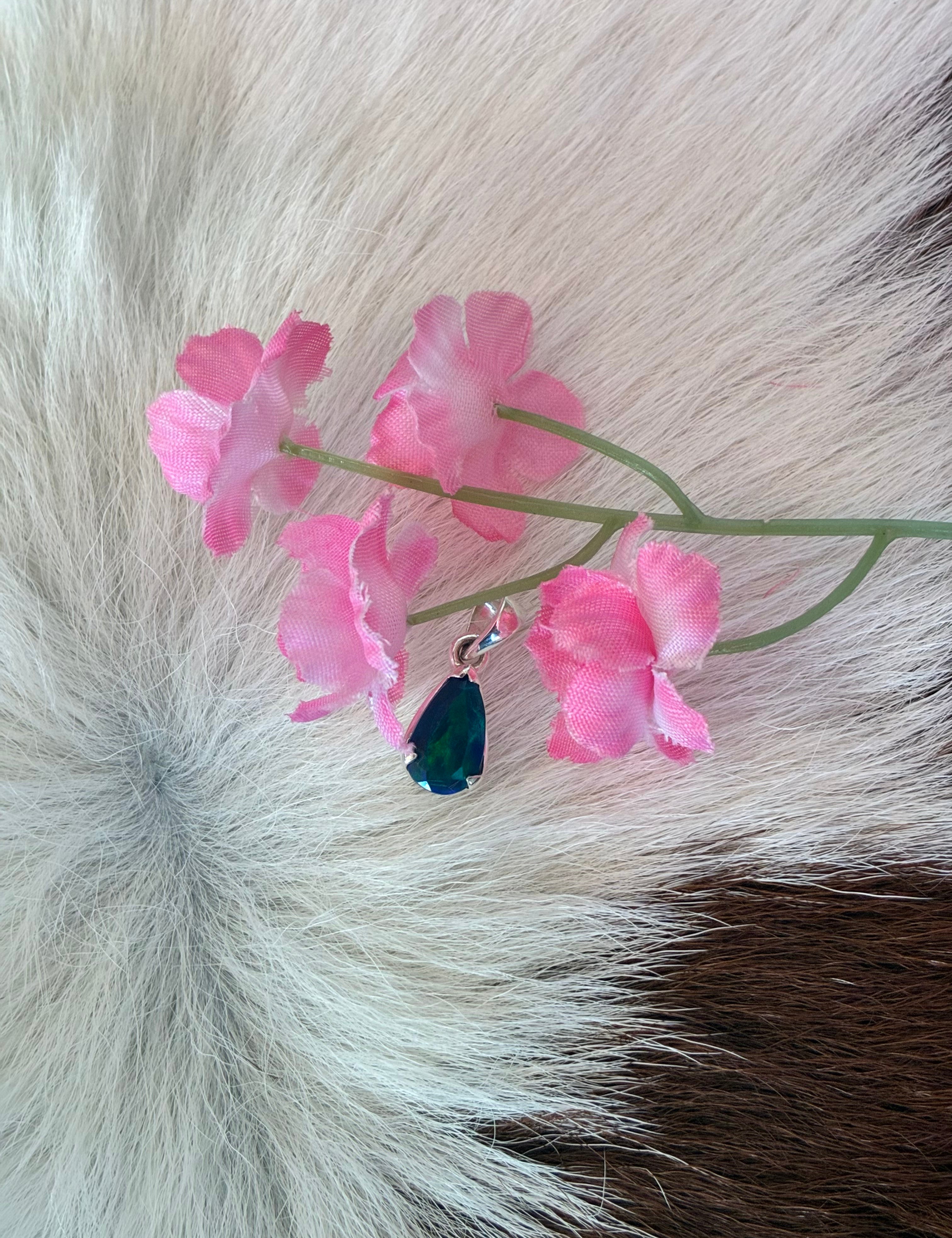 Black Ethiopian opal teardrop pendant in silver setting, displaying vibrant green and blue hues, surrounded by pink floral accents on a textured fur background.