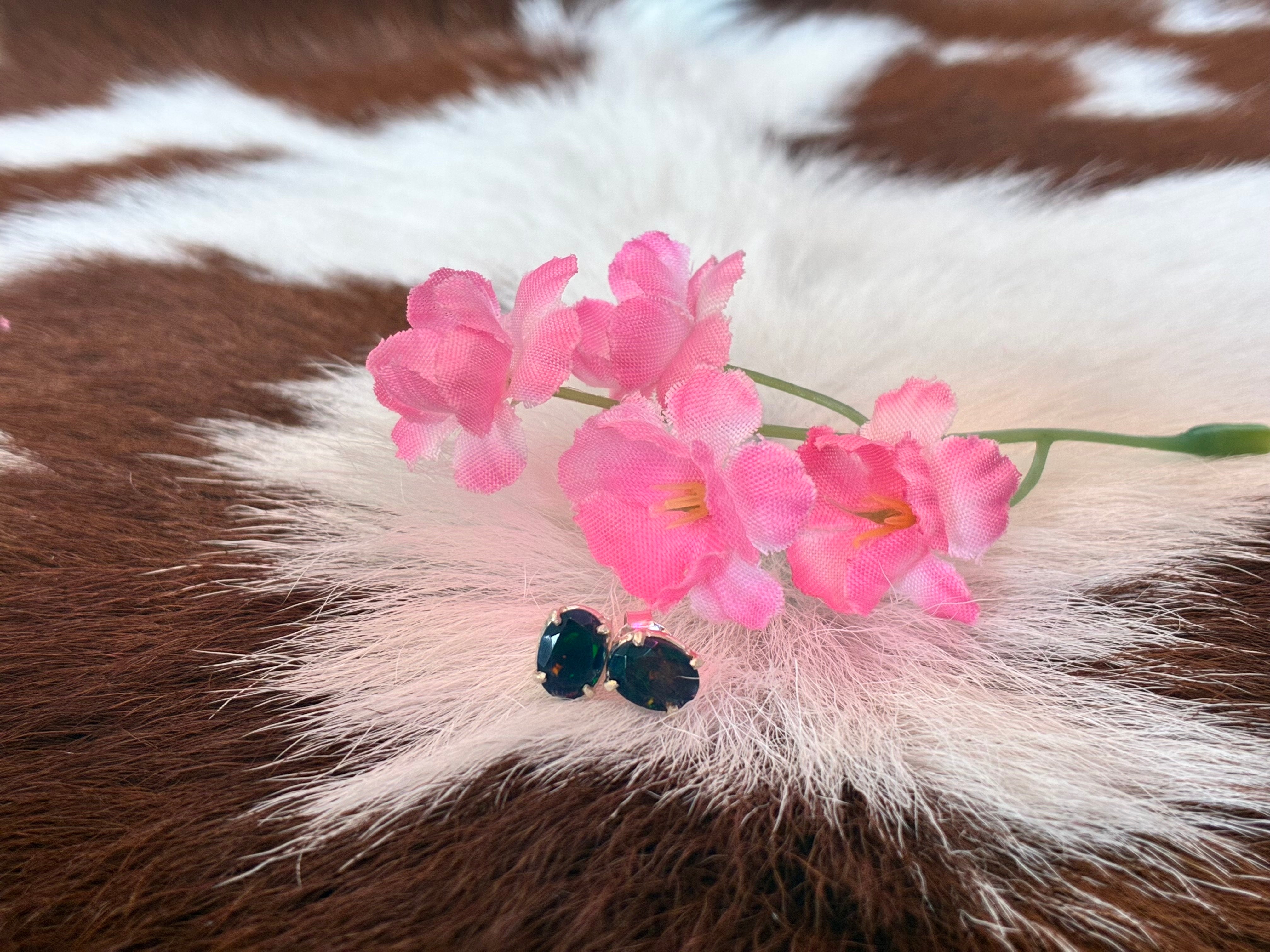 Black Ethiopian opal earrings with vibrant green and blue hues set in silver, surrounded by pink flowers on a textured fur background.Tags