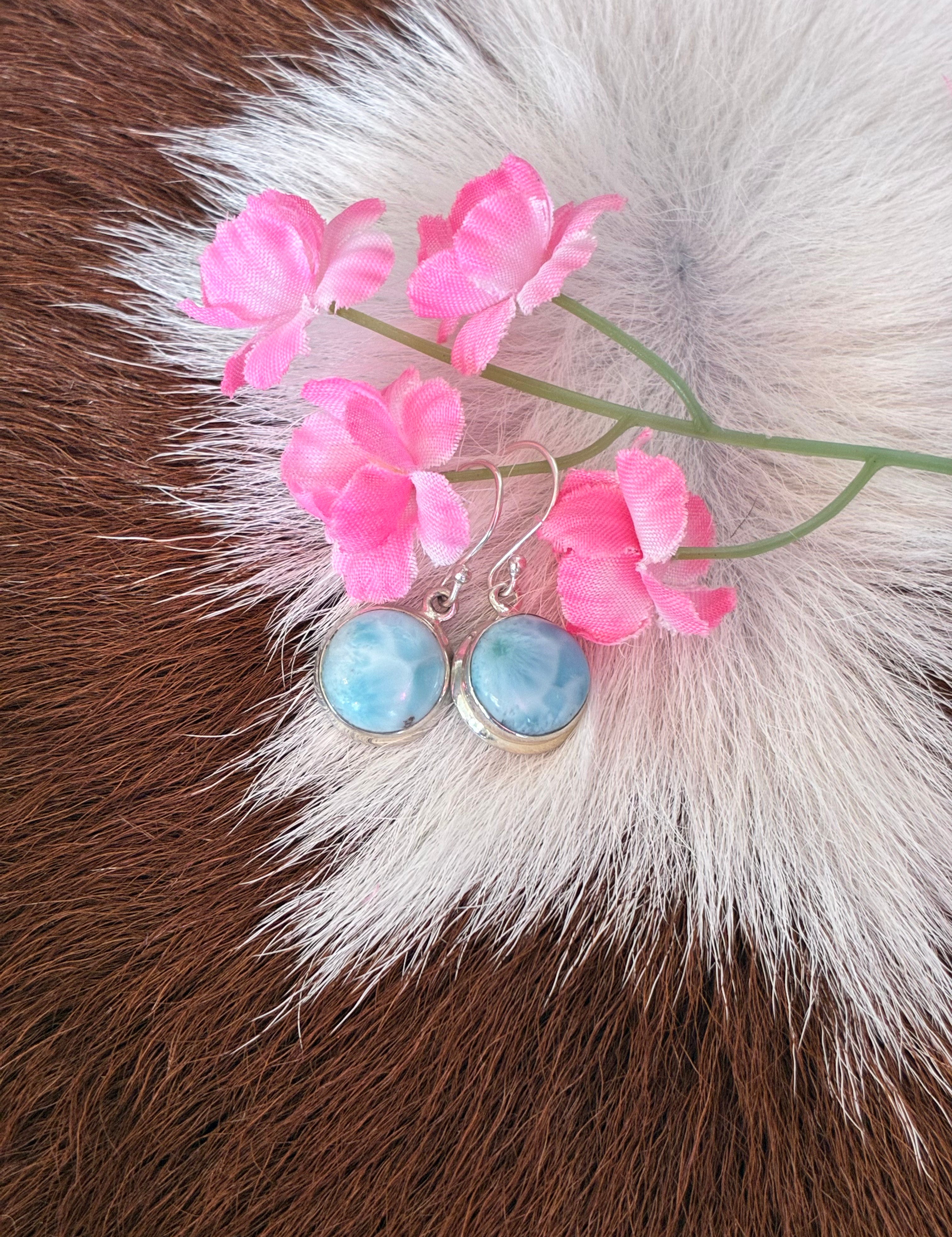 Round Larimar earrings featuring soft blue hues set in silver, surrounded by pink flowers on a textured fur background.
