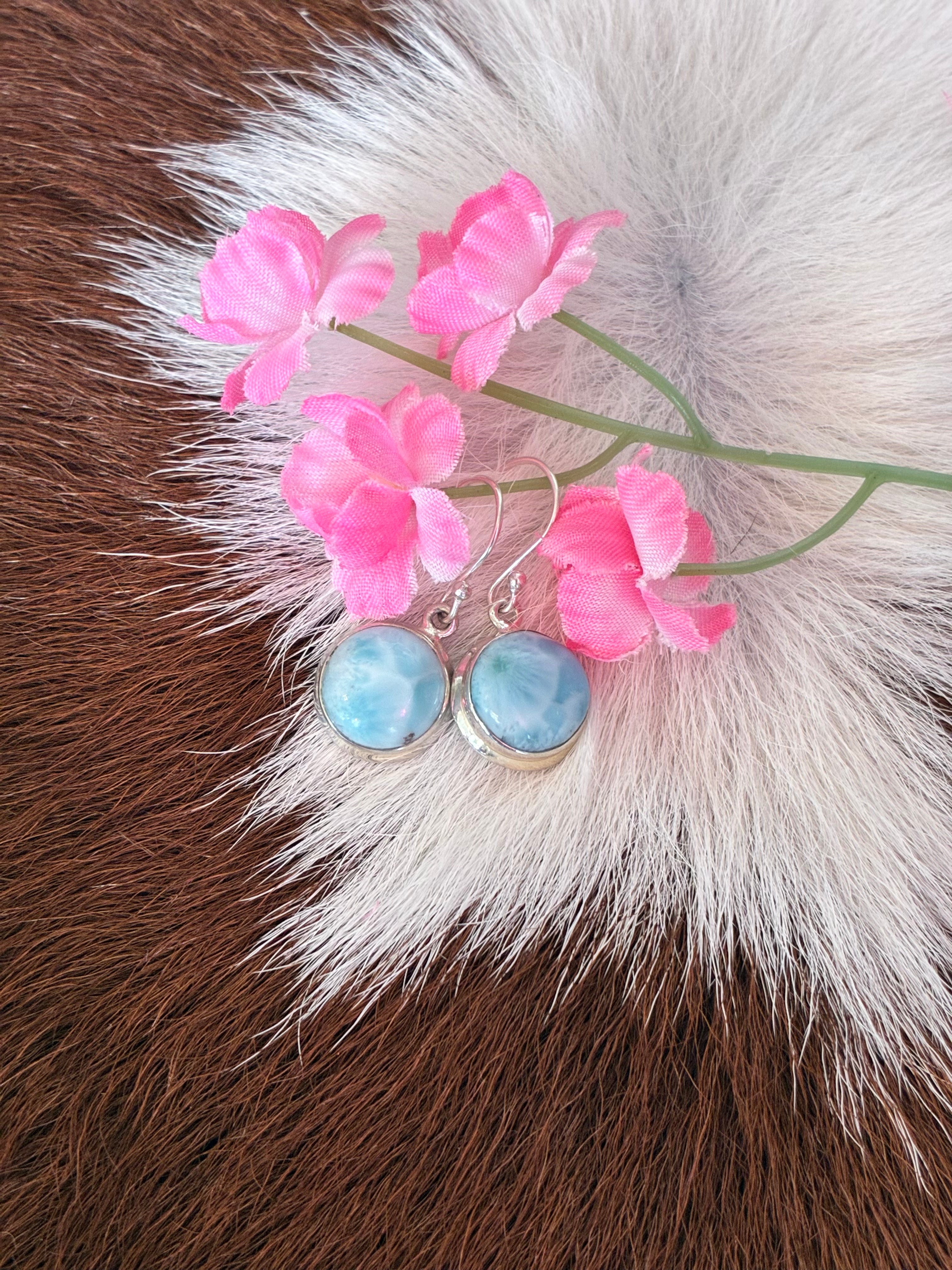 Round Larimar earrings featuring soft blue hues set in silver, surrounded by pink flowers on a textured fur background.