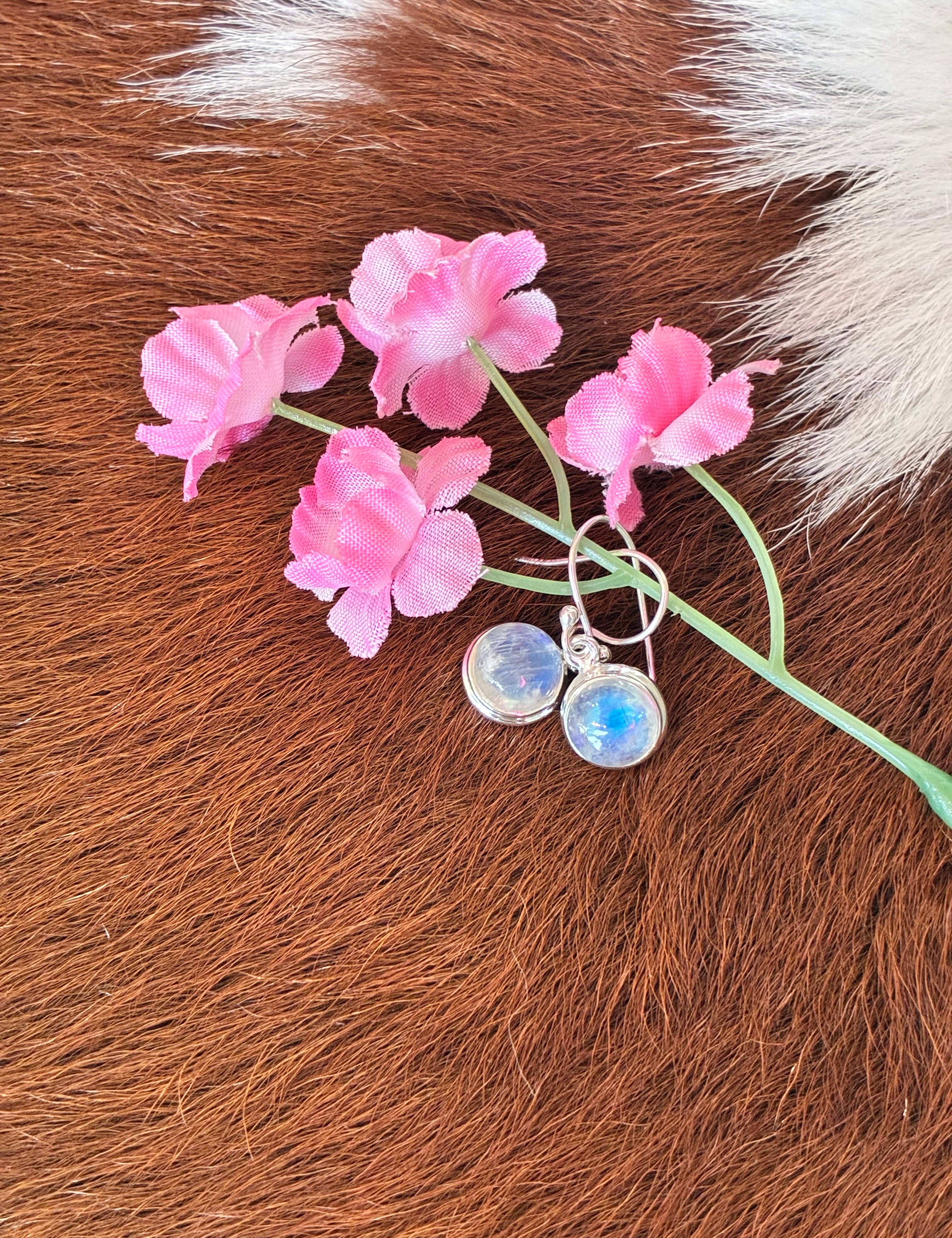 Round moonstone earrings featuring a captivating play of light set in silver, surrounded by pink flowers on a textured fur background.