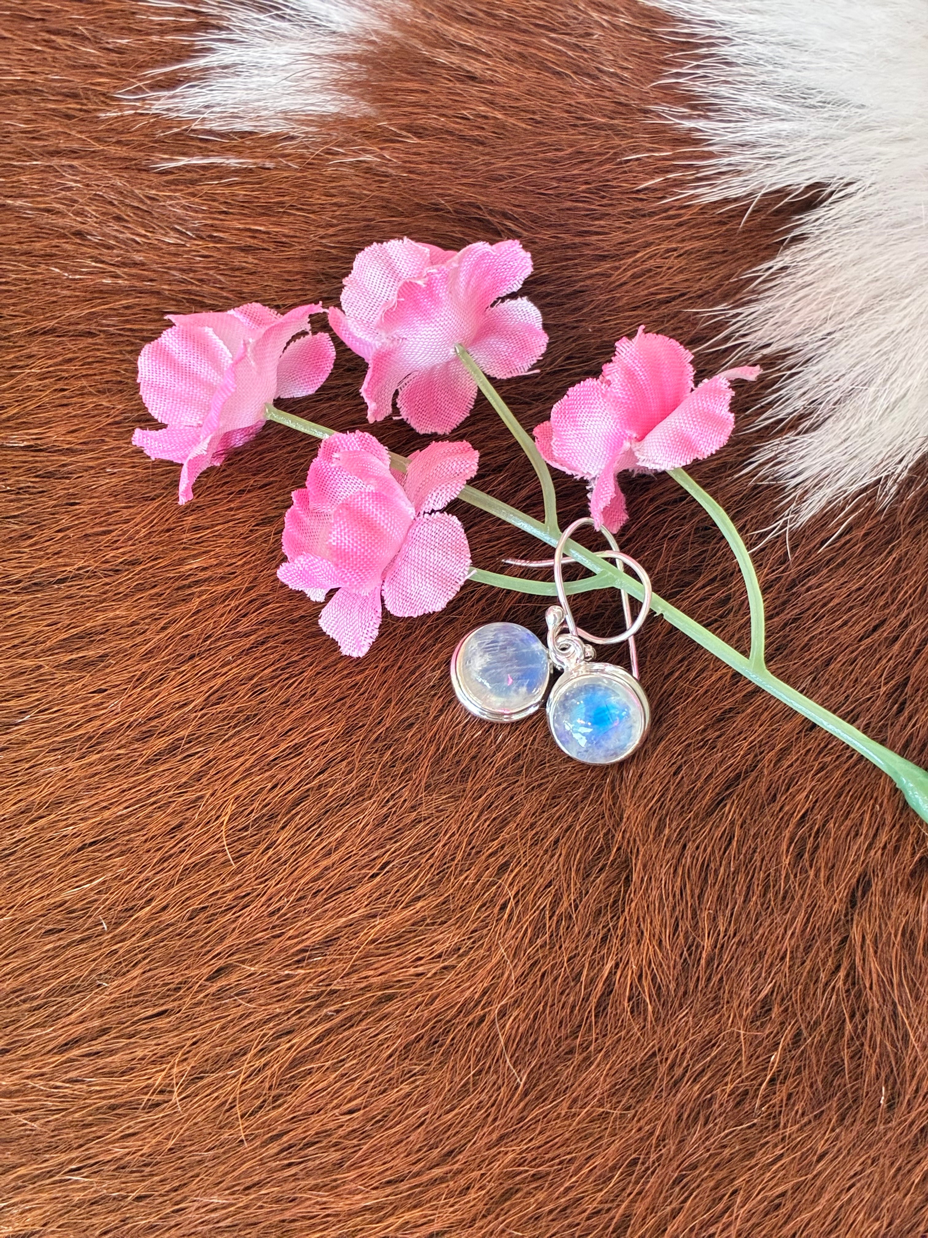 Round moonstone earrings featuring a captivating play of light set in silver, surrounded by pink flowers on a textured fur background.