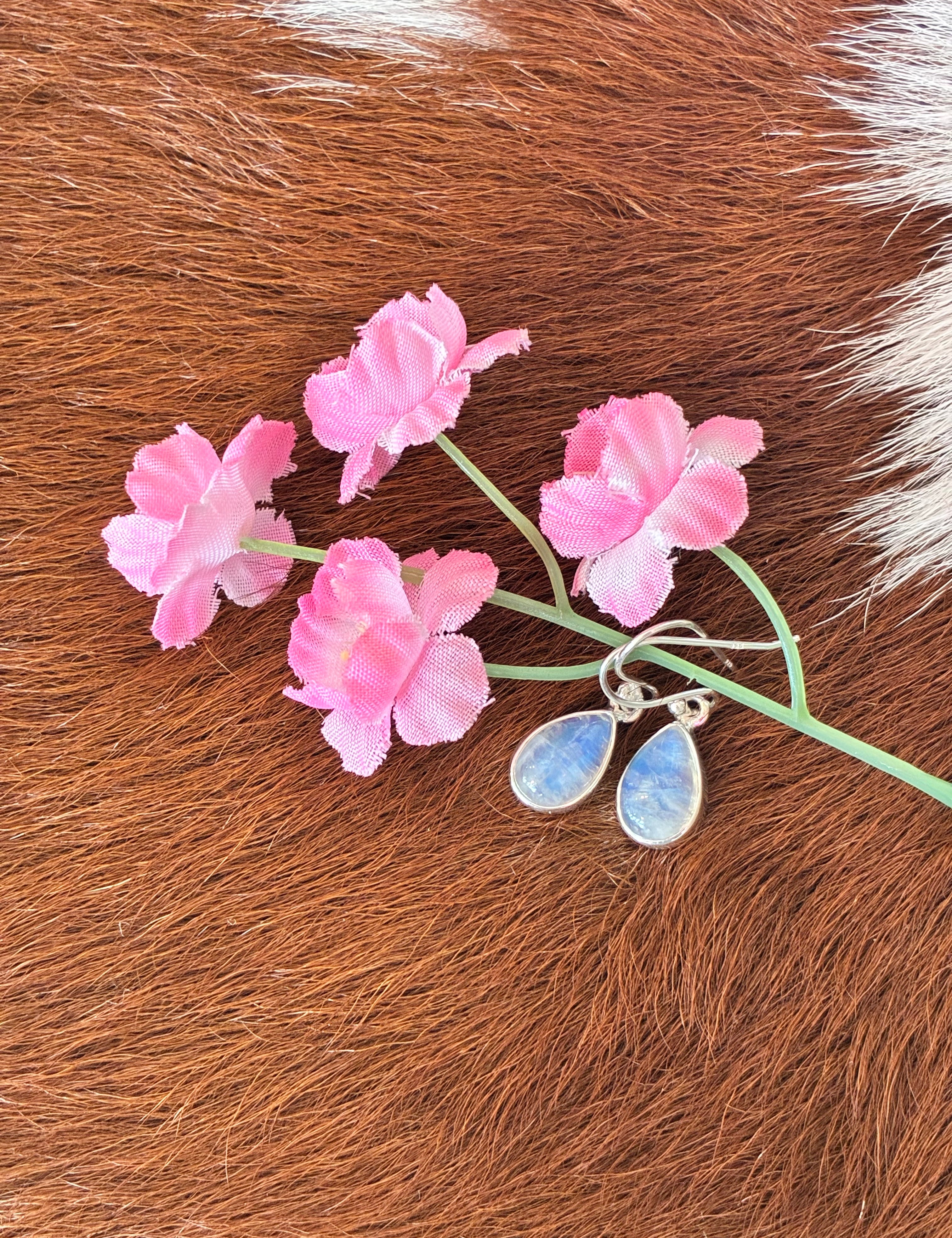 Teardrop moonstone earrings with soft blue and white hues set in silver, surrounded by pink flowers on a textured fur background.