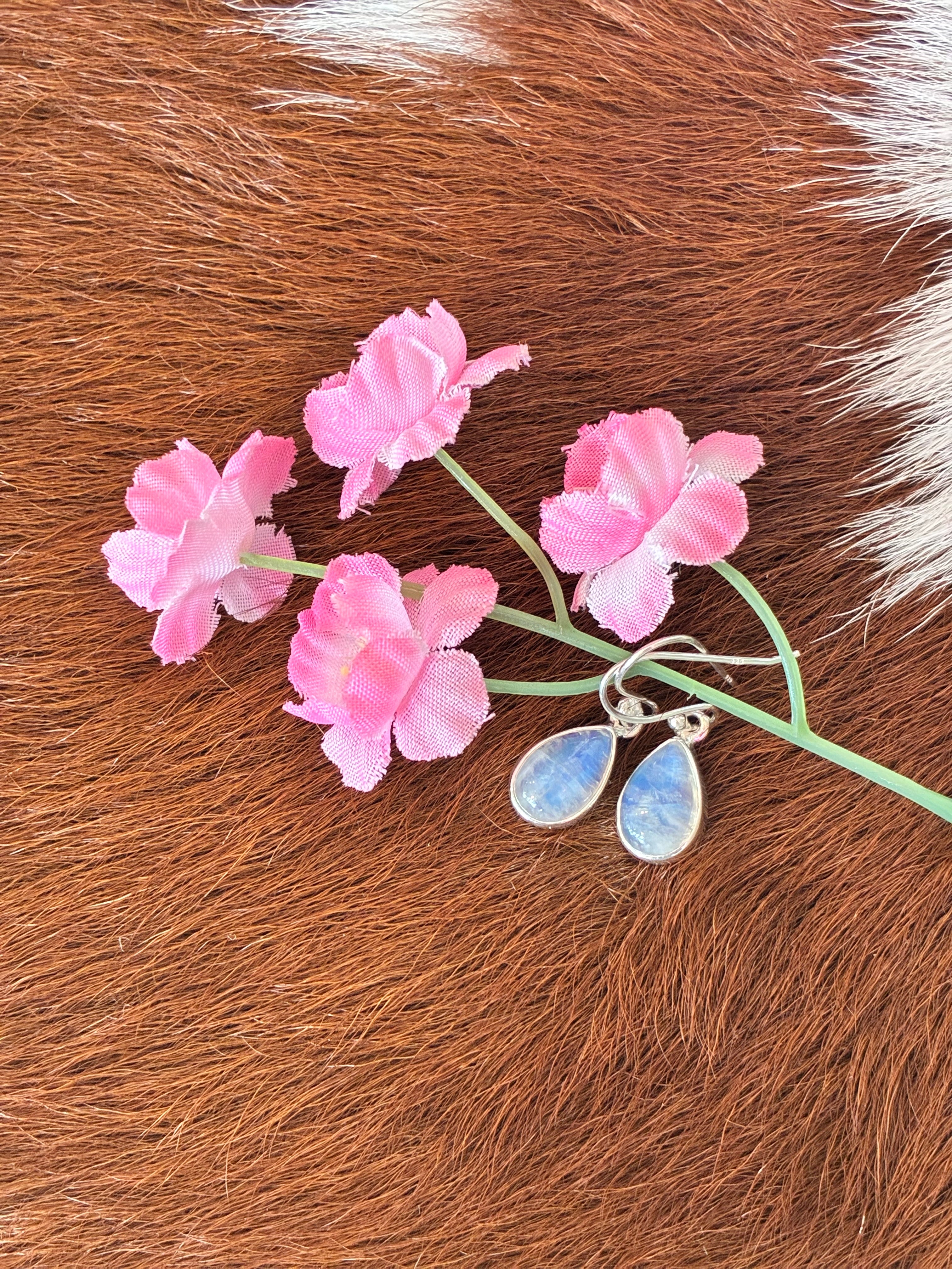 Teardrop moonstone earrings with soft blue and white hues set in silver, surrounded by pink flowers on a textured fur background.