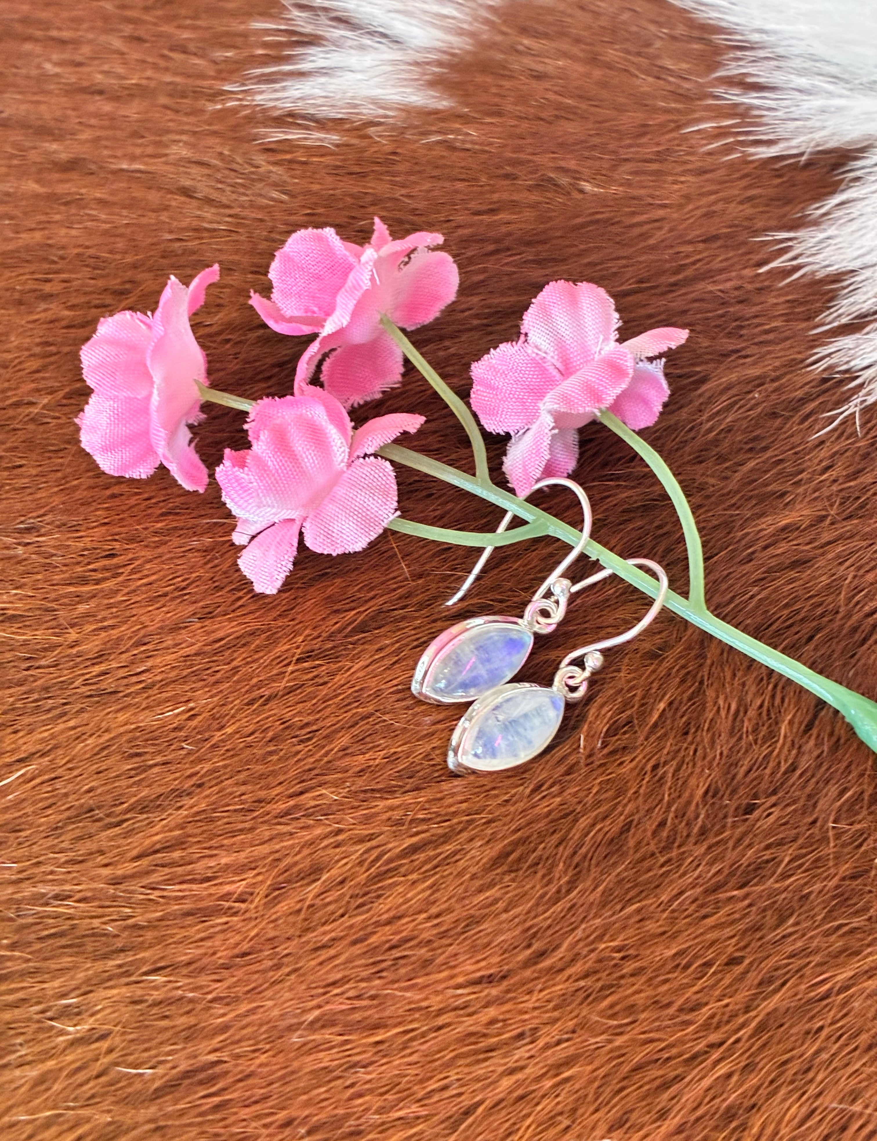 Marquise moonstone earrings with soft blue and pink hues set in silver, surrounded by pink flowers on a textured fur background.