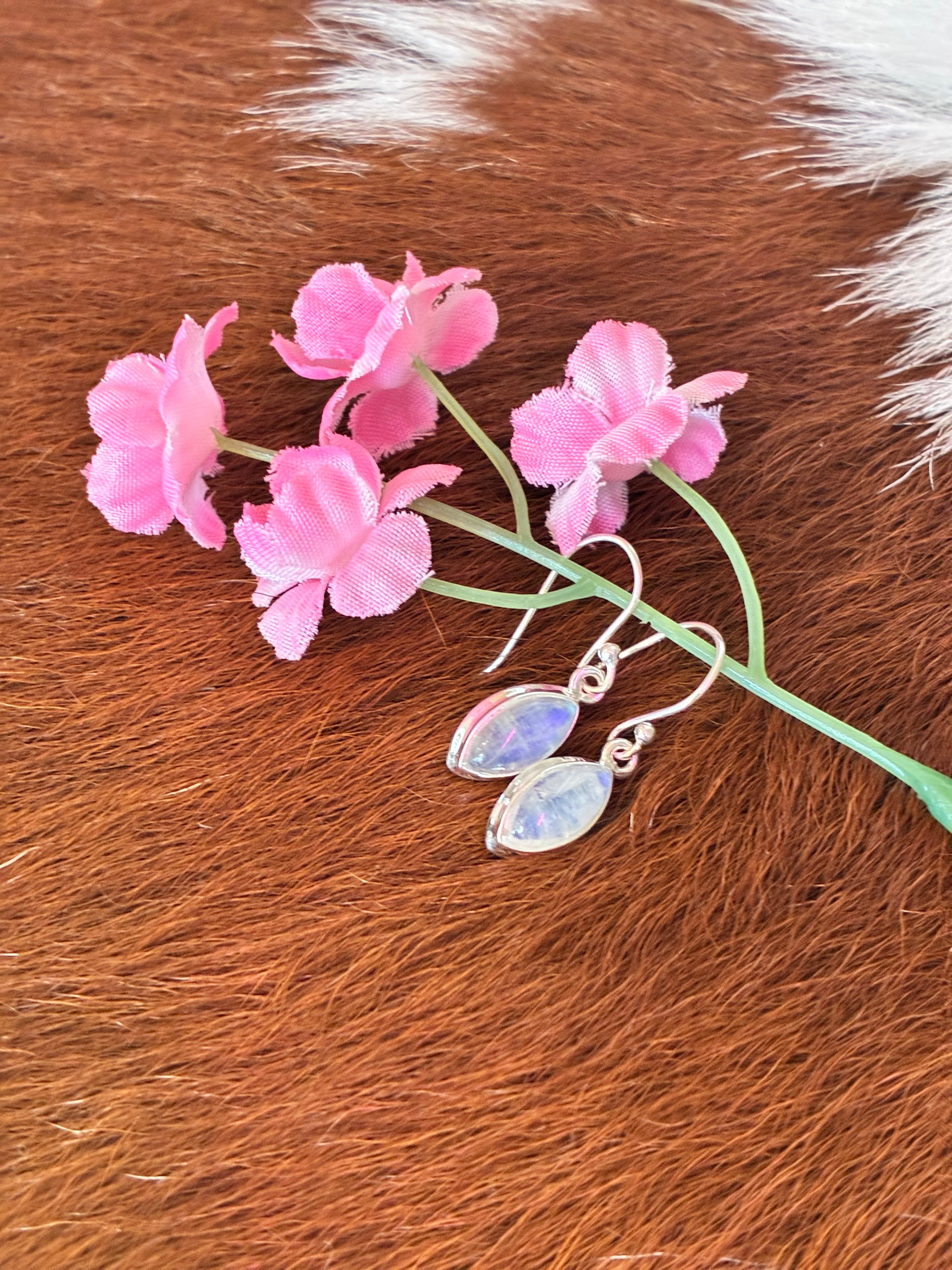 Marquise moonstone earrings with soft blue and pink hues set in silver, surrounded by pink flowers on a textured fur background.