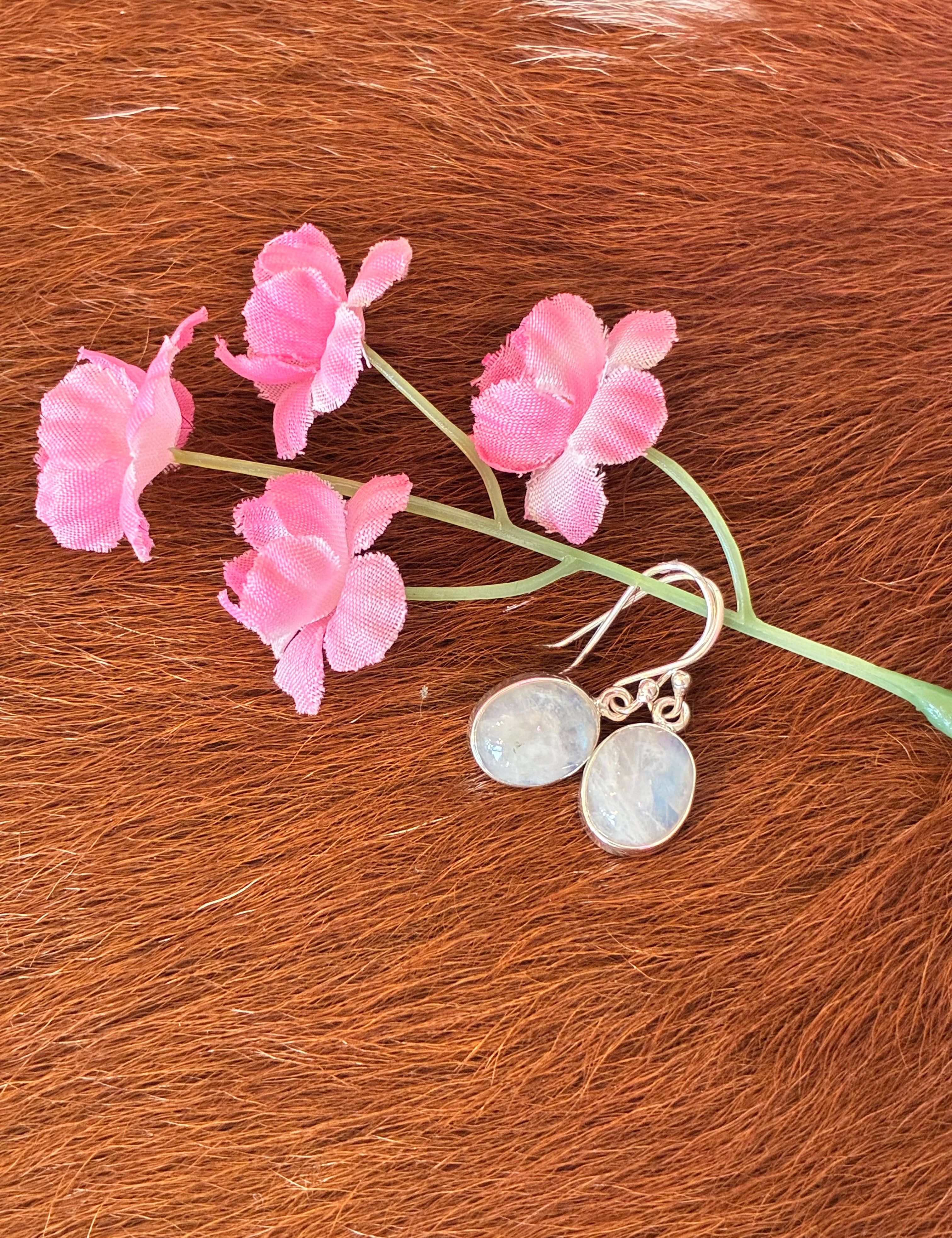 Oval moonstone earrings with soft blue and white hues set in silver, surrounded by pink flowers on a textured fur background.