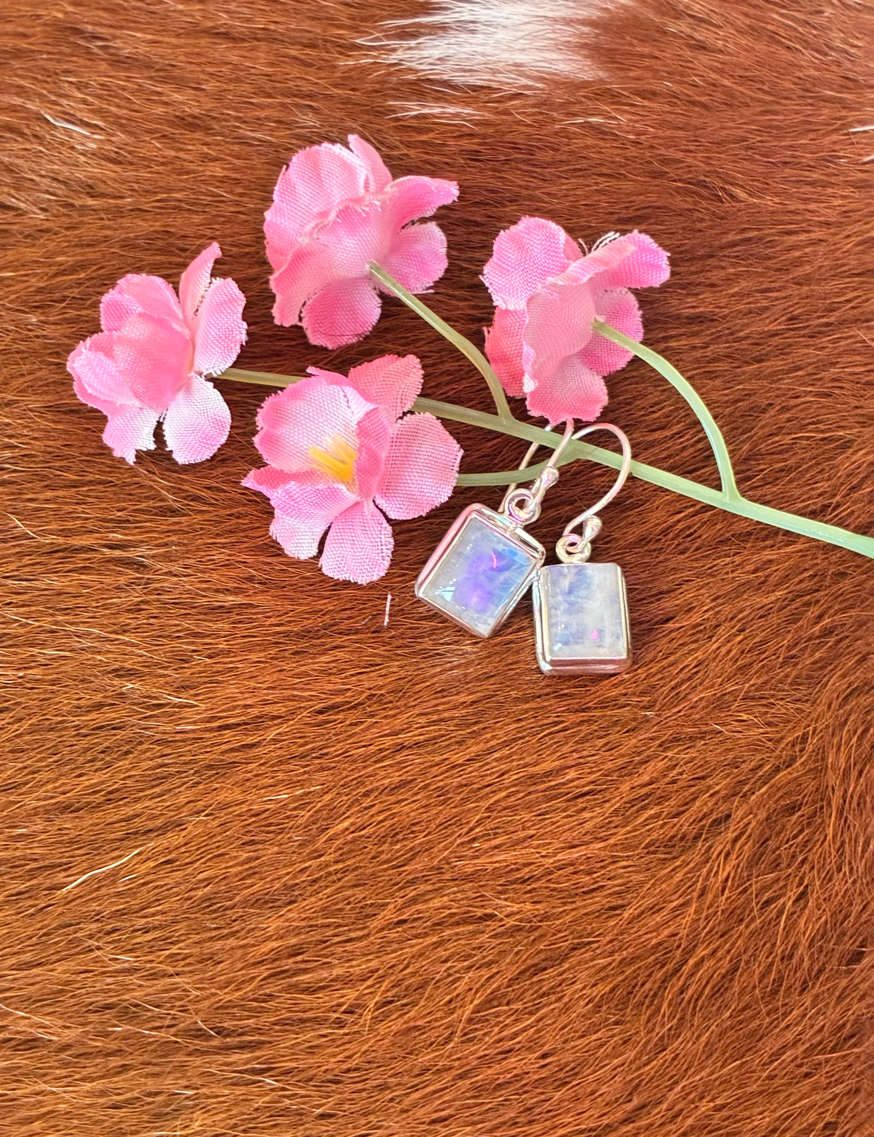 Rectangular moonstone earrings featuring a captivating play of colors set in silver, surrounded by pink flowers on a textured fur background.