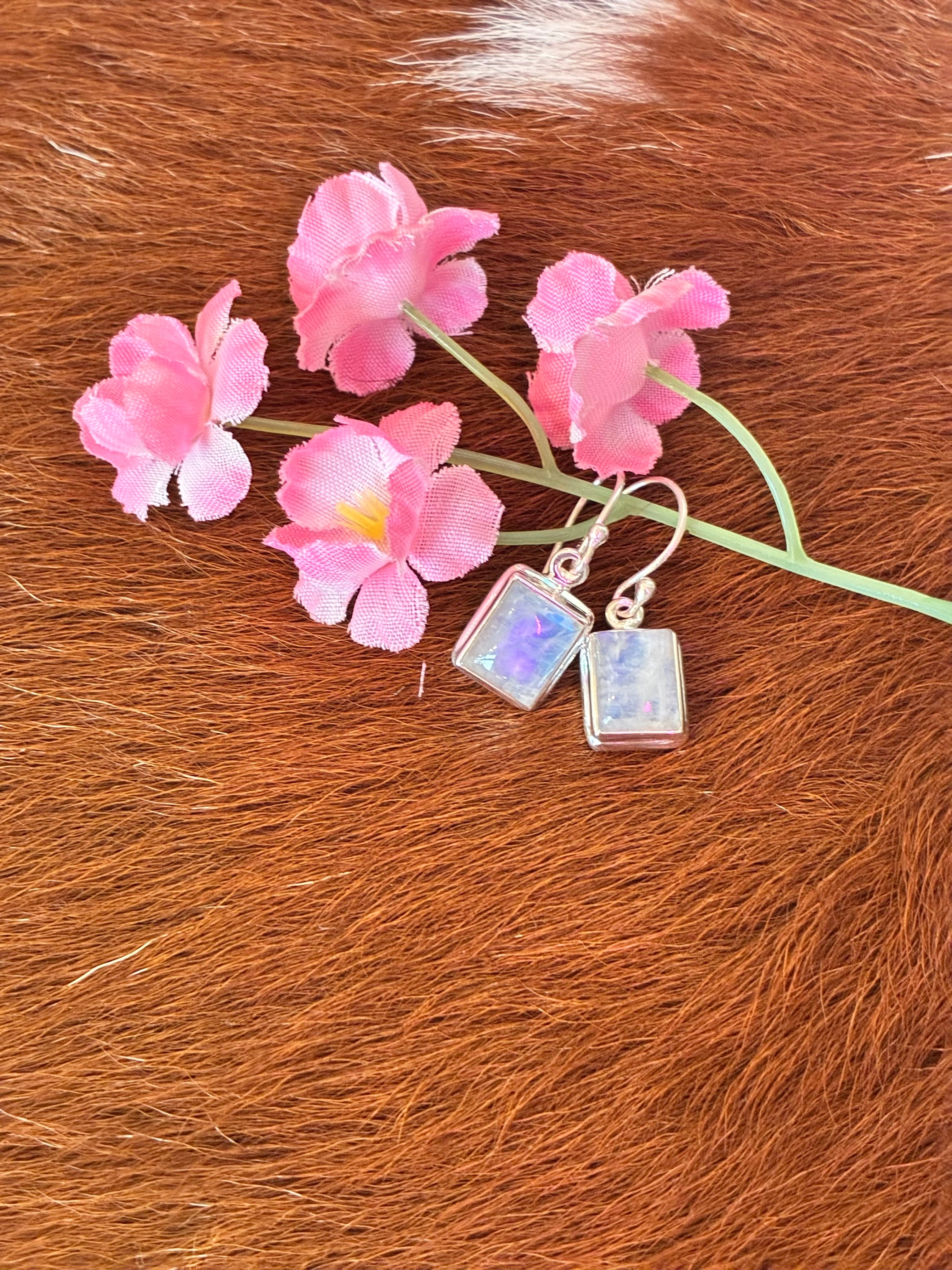 Rectangular moonstone earrings featuring a captivating play of colors set in silver, surrounded by pink flowers on a textured fur background.