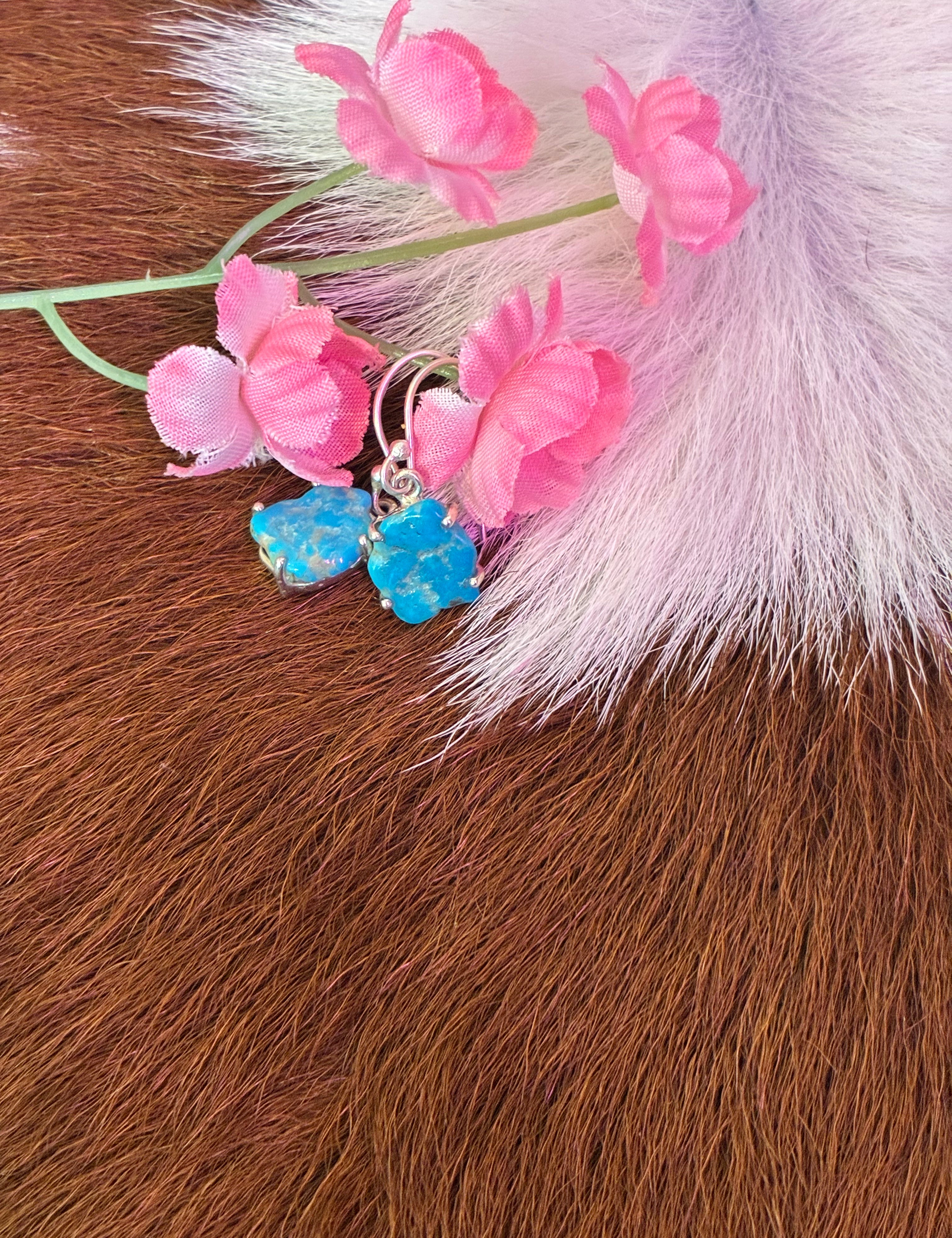 Organic-shaped turquoise earrings set in silver, surrounded by pink flowers on a textured fur background.