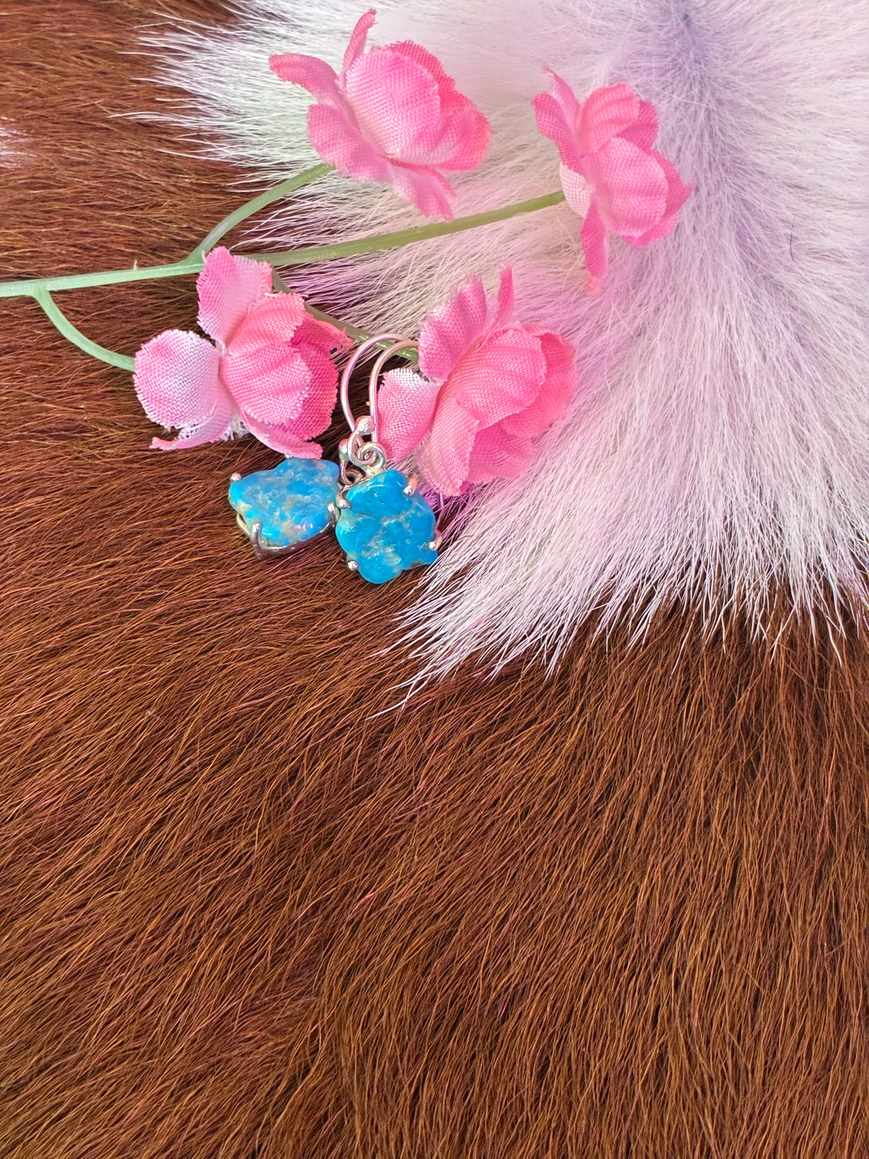 Organic-shaped turquoise earrings set in silver, surrounded by pink flowers on a textured fur background.