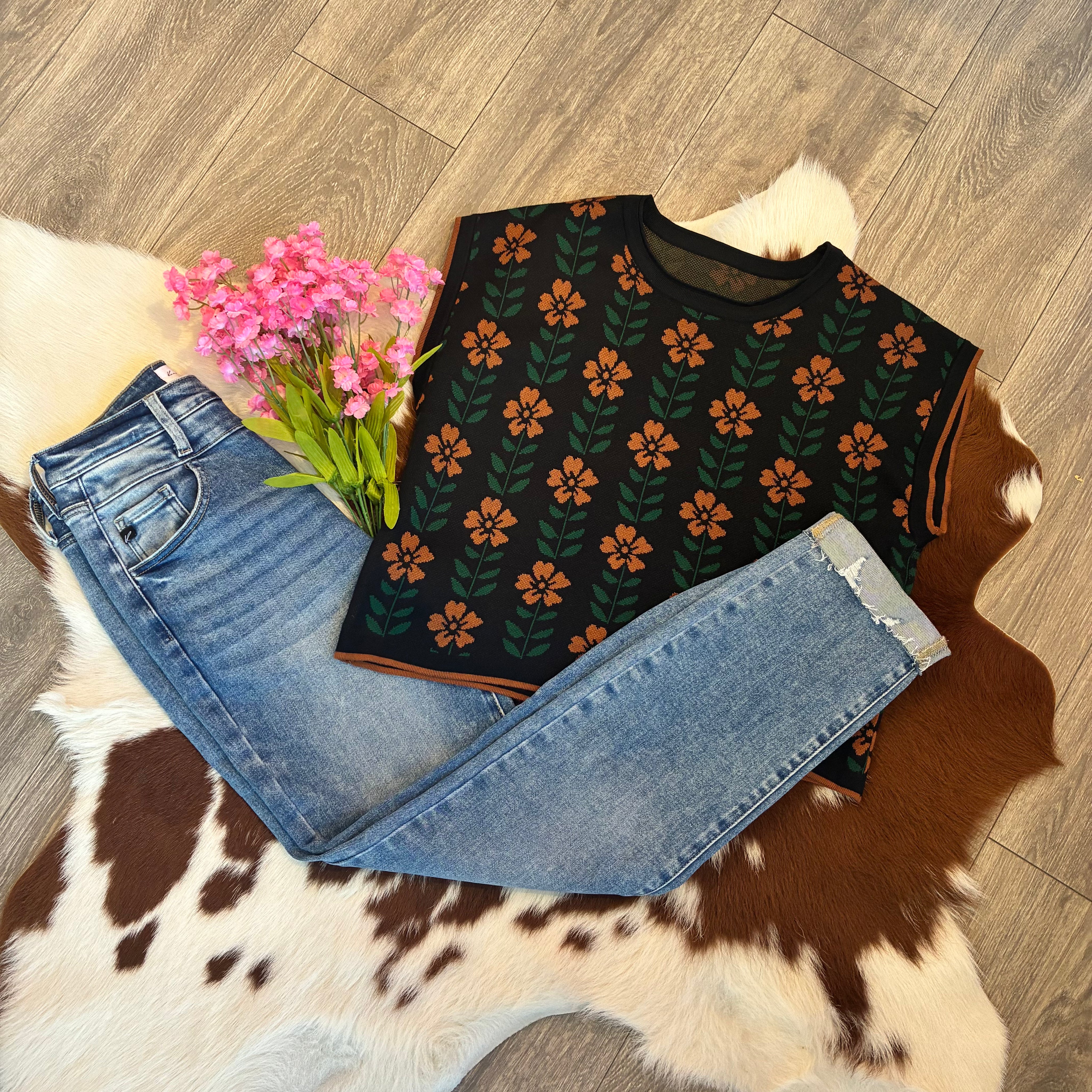 Black sleeveless top with floral pattern, blue jeans, and pink flowers on a wooden floor.