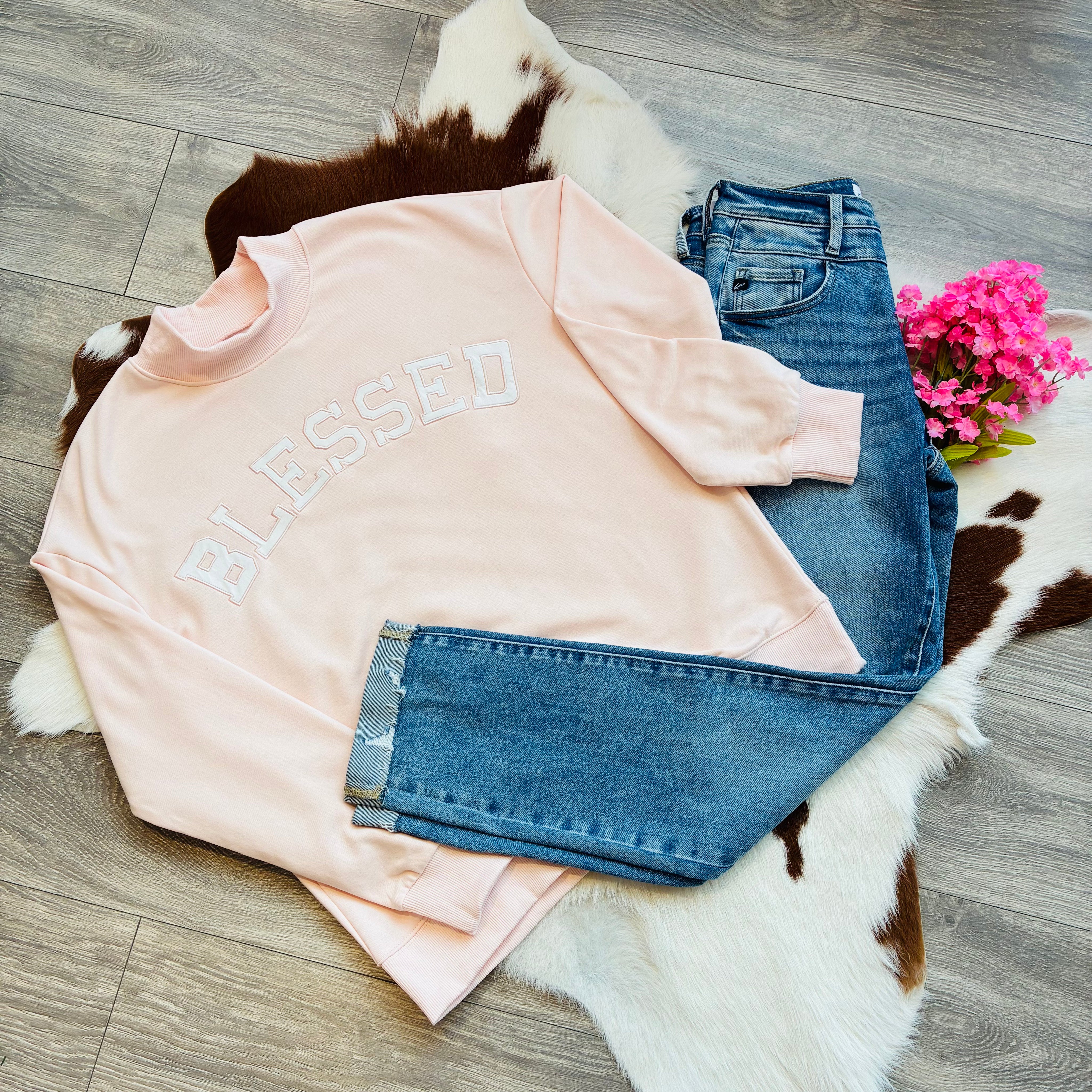 Pink sweatshirt with 'BLESSED' text and blue jeans on a fur rug with flowers.
