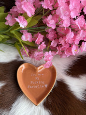 A heart-shaped ceramic dish in a warm tan color with a glossy finish sits on a brown and white cowhide surface. The dish features the phrase "You are My F***ing Favorite ♥" in white text. Above the dish, soft pink artificial flowers with green leaves add a delicate contrast to the rustic background.