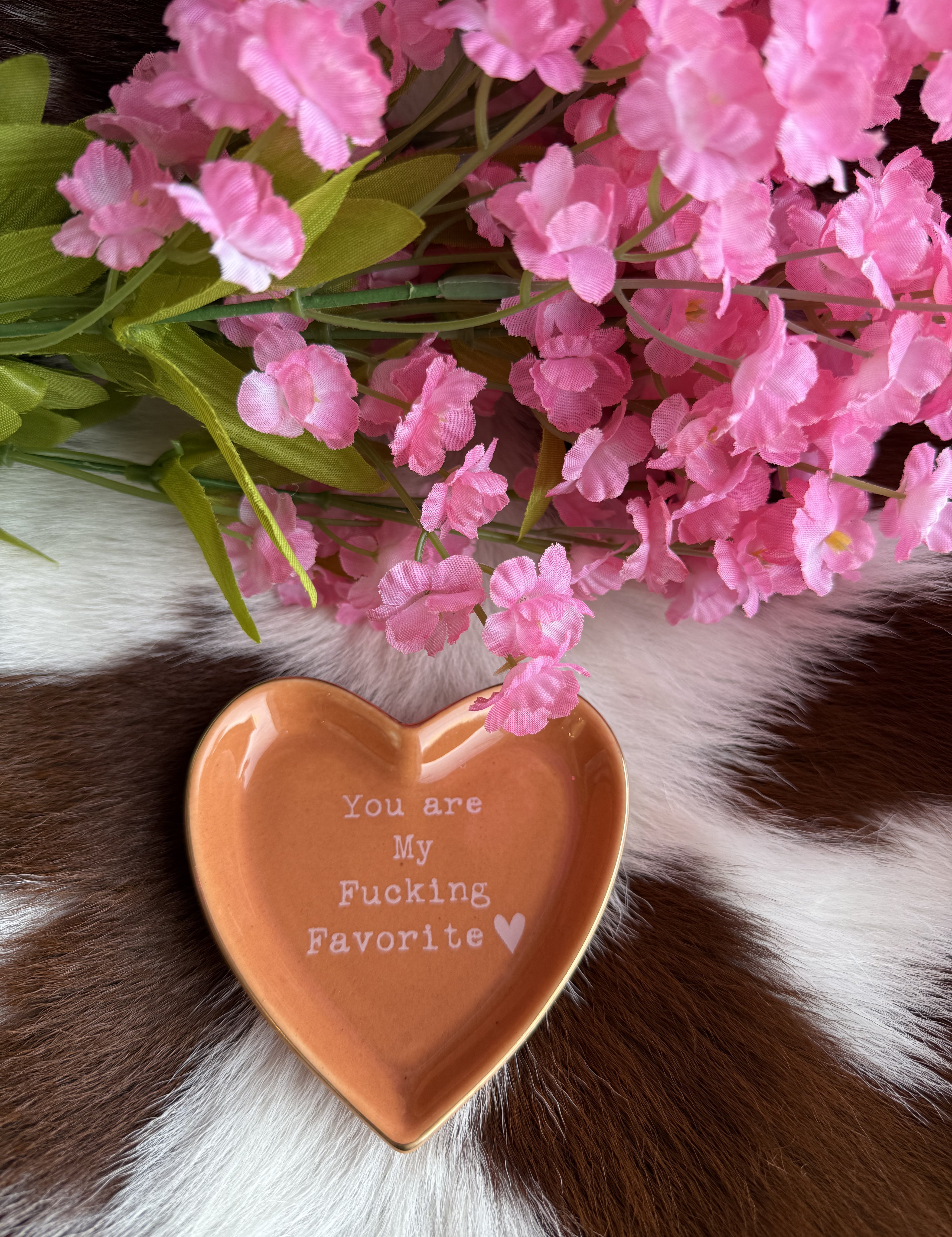 A heart-shaped ceramic dish in a warm tan color with a glossy finish sits on a brown and white cowhide surface. The dish features the phrase "You are My F***ing Favorite ♥" in white text. Above the dish, soft pink artificial flowers with green leaves add a delicate contrast to the rustic background.