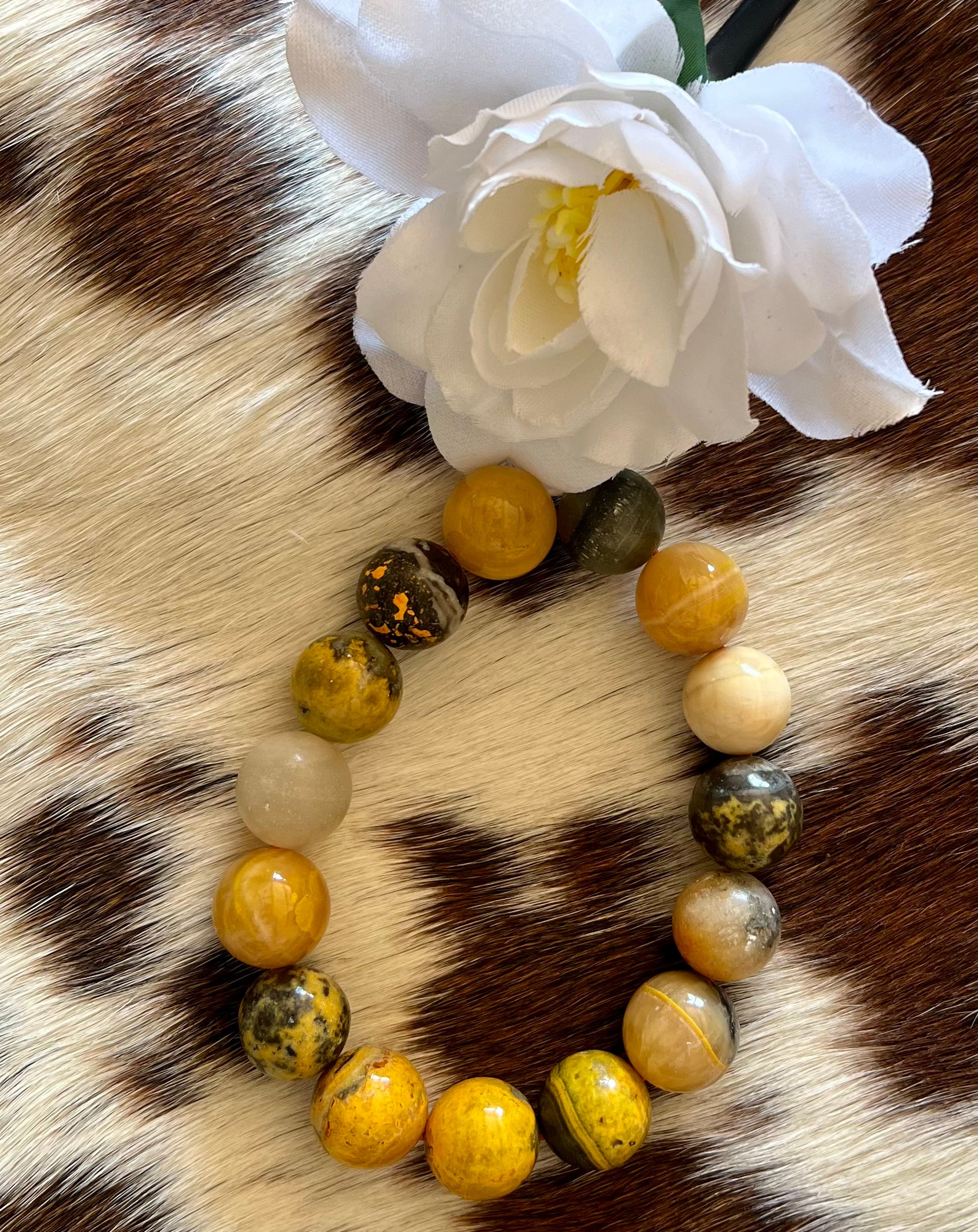 A close-up of a beaded bracelet featuring round, polished stones in shades of yellow, gold, beige, and black with unique marbled patterns. The bracelet is displayed on a cowhide surface with a mix of brown and white fur, adding a rustic and Western-inspired aesthetic. A delicate white fabric flower is positioned near the top, enhancing the elegant and earthy feel of the image.