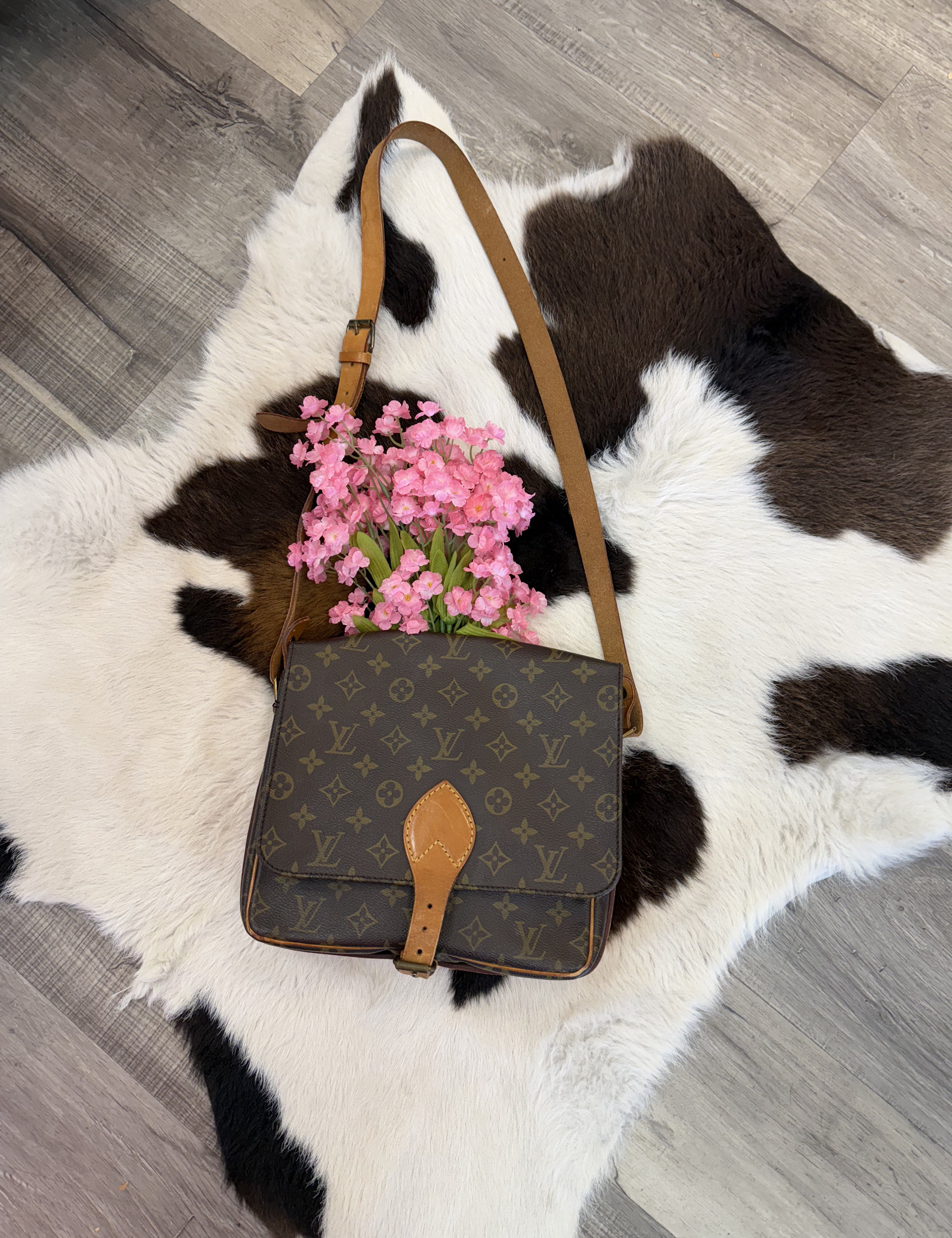 A vintage designer crossbody bag with a monogram pattern and tan leather strap displayed on a black and white cowhide rug. The bag features a buckle closure with a leather accent. A bouquet of pink flowers is tucked inside, adding a charming and feminine touch. The display is set on a wood floor, enhancing the rustic yet elegant aesthetic