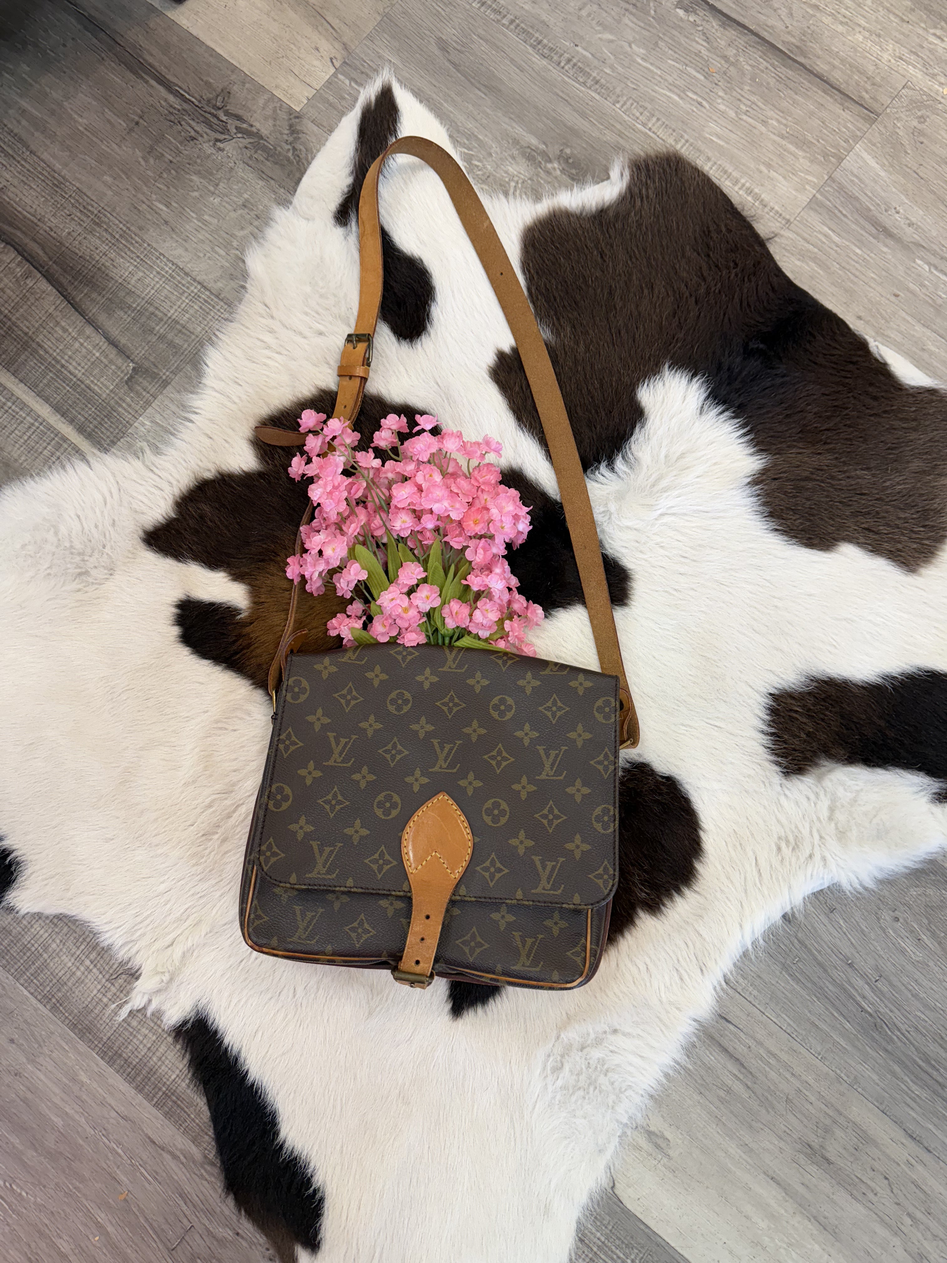 A vintage designer crossbody bag with a monogram pattern and tan leather strap displayed on a black and white cowhide rug. The bag features a buckle closure with a leather accent. A bouquet of pink flowers is tucked inside, adding a charming and feminine touch. The display is set on a wood floor, enhancing the rustic yet elegant aesthetic