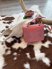 A pink embossed leather handbag with intricate floral tooling rests against a white cow skull adorned with artificial succulents. The bag has gold-toned metal rings attaching the handle and a matching pink crossbody strap. The background features a cowhide rug with brown and white patches, complementing the rustic, Western-inspired aesthetic